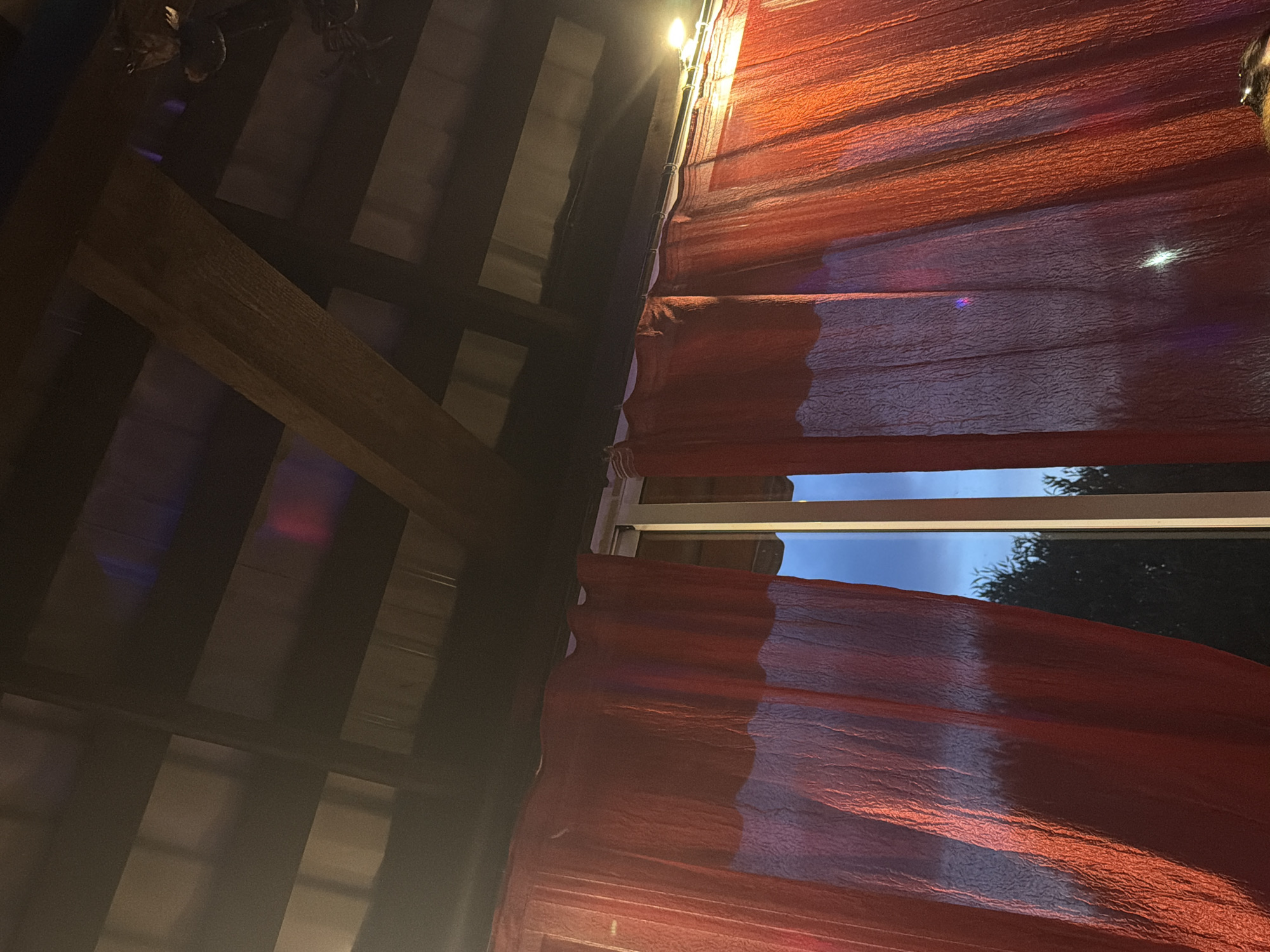 The image shows a view from under a covered patio or porch looking out at the night sky and trees. Red curtains are drawn, revealing a narrow opening through which the outside is visible. The sky is a deep blue, indicating it is nighttime, and silhouetted against it are the dark shapes of trees. The roof structure above is made of dark wood with exposed beams and has a corrugated metal ceiling. Lights are visible, one in the upper right corner, and there are colored reflections on the roof and curtains, suggesting some form of ambient lighting. The scene appears to be outdoors, possibly a restaurant or a residential patio, in Chaikovsky, Russia, given the request.