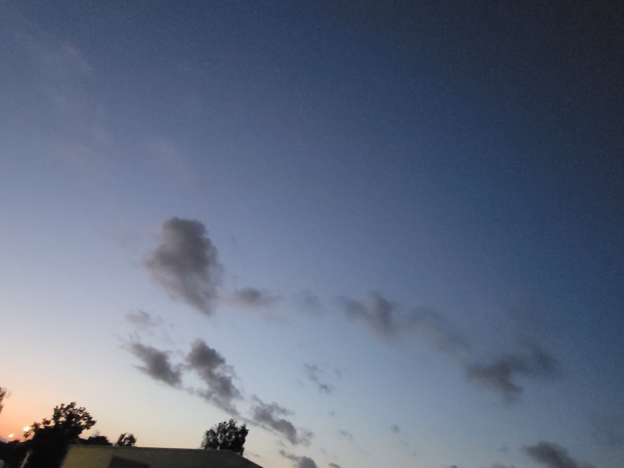 This image depicts a twilight sky over a silhouetted landscape, likely in Ozolnieki, Latvia. The sky transitions from a pale orange glow near the horizon on the left to a deep blue overhead. Wispy, dark clouds are scattered across the sky, some appearing as elongated streaks and others as more amorphous shapes. In the lower portion of the image, the dark silhouettes of trees and the top of a building are visible against the lighter sky. The overall scene conveys a serene, end-of-day atmosphere. There are no visible people or specific actions taking place.