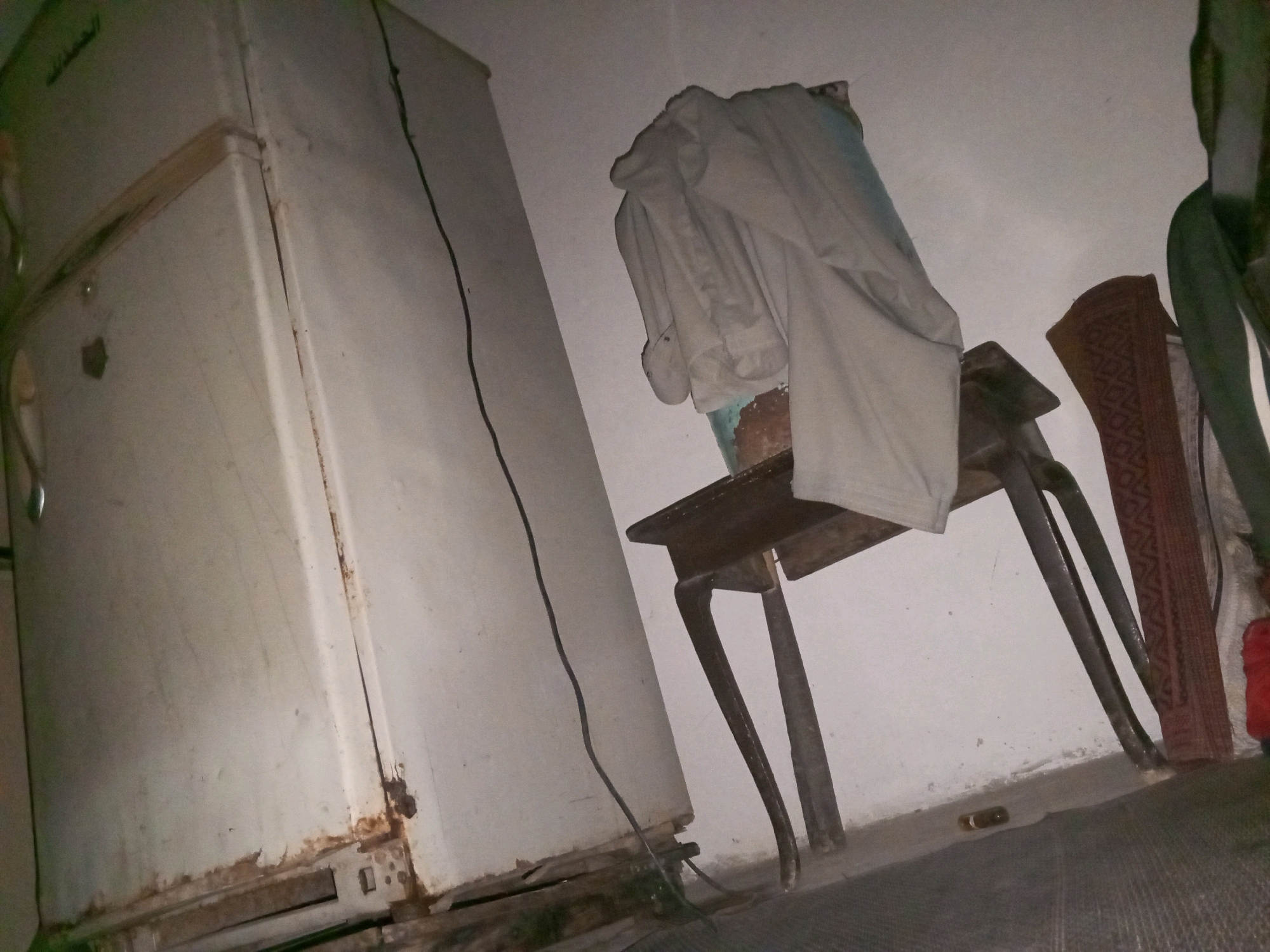 The image depicts an indoor scene, likely a room in a dwelling located in Kanju, Pakistan. The primary focus is a large, off-white refrigerator on the left side of the frame, showing signs of rust and wear, particularly around its base. A black electrical cord runs from the refrigerator, extending vertically along the wall. To the right of the refrigerator, a small, dark wooden table with intricate carvings on its legs is positioned. Draped over the table are several pieces of light-colored clothing. Behind the table, a woven mat with a reddish-brown geometric pattern is visible, leaning against the wall. The wall itself is plain and appears to be painted a light color. The floor is a dark, textured surface. The lighting is dim, suggesting the photograph was taken in low-light conditions, possibly at night or in an area with limited natural light. There are no people visible in the image, and no specific actions are being performed. The environment appears to be a somewhat cluttered and utilitarian living space.
