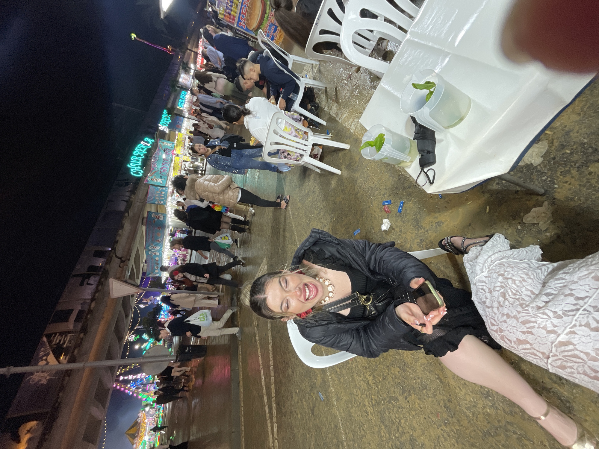 The image captures an outdoor scene at night, likely in Jerez De La Frontera, Spain, given the signage visible in the background. A woman is seated in a white plastic chair, smiling broadly at the camera. She wears a black jacket, a dark top, a pearl necklace, and holds a smartphone. To her right, another person is seated, wearing a white lace dress and gold heels. The ground appears wet, and there are discarded items scattered around. In the background, there is a brightly lit fairground with numerous stalls and a crowd of people. There is a table with water filled cups with leaves next to them and a black face mask.