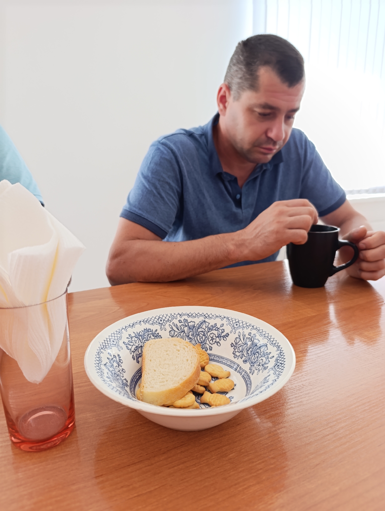 A man in a blue polo shirt is sitting at a wooden table, looking down at a black mug he is holding. His right hand is holding the mug, and his left hand is stirring the contents of the mug with a utensil. In front of him is a white bowl with a blue floral pattern. The bowl contains a slice of bread and several small, fish-shaped crackers. To the left of the bowl is a clear glass with white napkins. The background is a white wall with vertical blinds on the right side. The scene appears to be indoors, possibly in a dining area or cafe. The overall lighting suggests it is daytime.
