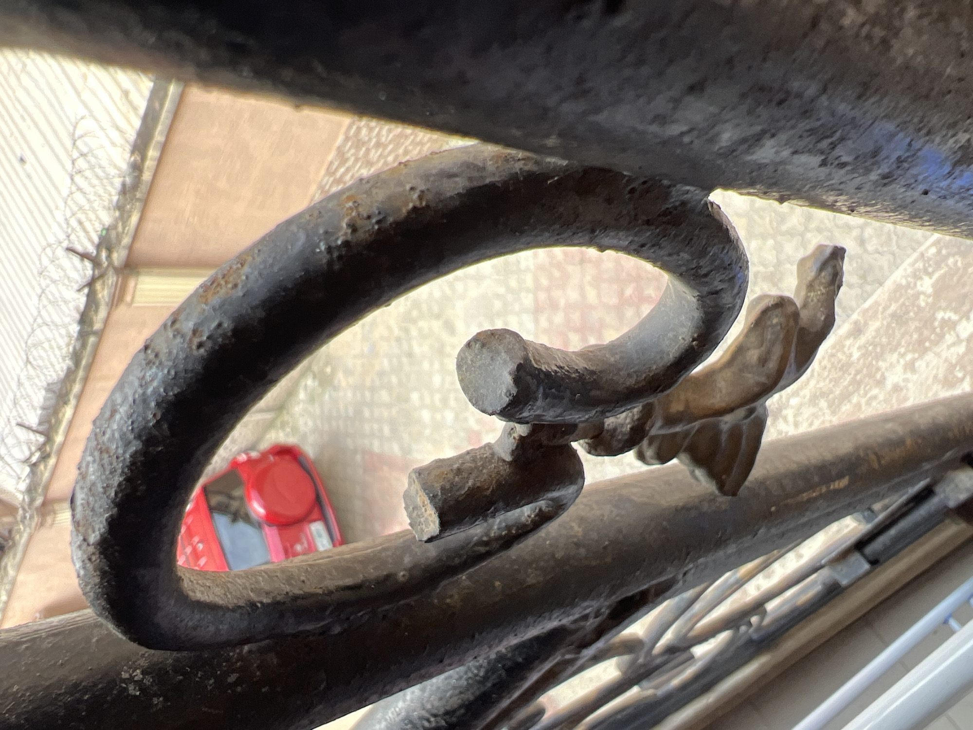 The media file is a close-up, overhead shot of weathered, dark metal railings, likely from a balcony or staircase. The railings are curved and show signs of rust and age. Through the gaps in the railings, a glimpse of the environment below is visible.
The scene below appears to be an outdoor area with a textured, light-colored ground, possibly a paved courtyard or walkway. To the left, a barbed wire fence is visible, suggesting a secure or restricted area. In the mid-ground, a bright red, small vehicle, resembling a car or a utility vehicle, is parked. Its reflective surfaces are visible, indicating it's a modern vehicle.
The lighting suggests it is daytime, with sunlight casting shadows and highlighting the textures of the metal. The overall impression is one of urban infrastructure, possibly in a residential or commercial setting in Lagos, Nigeria, given the contextual information. There is no visible text in the image. The focus is on the weathered metal and the partially obscured view of the surroundings, creating a sense of depth and hinting at life beyond the immediate foreground. No people are visible in the media file.