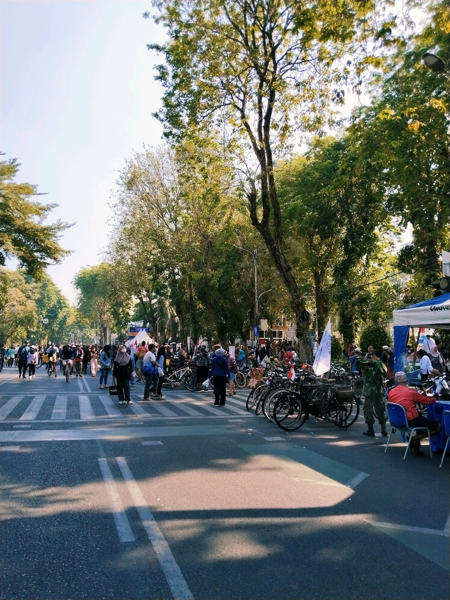 The image depicts a wide street scene in Surabaya, Indonesia, likely during a daytime event. A large crowd of people is gathered, many of them walking across a pedestrian crossing. Bicycles are parked and visible in groups on the side of the road. Several trees line the street, providing shade. A blue and white tent with the word "Universitas" printed on it is set up on the right side of the image, suggesting an event related to a university. People are dressed in casual attire, and some are wearing masks. The overall atmosphere appears to be lively and communal, with individuals engaged in various activities, from walking and cycling to interacting near the tent. The presence of a military-clad individual suggests a potential security presence or participation in the event. The lighting indicates it is a sunny day.