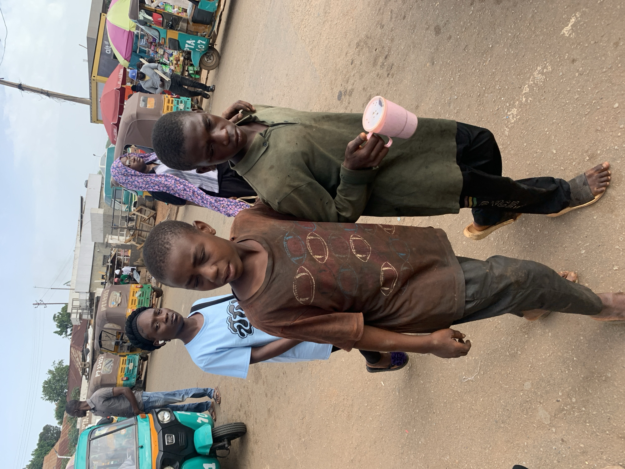 The image depicts a street scene in Gura Top, Nigeria, with several people and motorized rickshaws (also known as "keke") present. In the foreground, two young males are walking. The one on the left wears a brown t-shirt with a football-themed print and dark pants. The male on the right is wearing a green long-sleeved collared shirt and black pants, holding a pink cup. Behind them, a young female in a light blue t-shirt and black headscarf is visible. Further in the background, a woman in a colorful headscarf and glasses is seen, along with a man in a grey shirt and jeans. Several keke are parked or moving in the background, some with passengers. The environment appears to be a dusty road with buildings and stalls lining the sides. The sky is overcast.