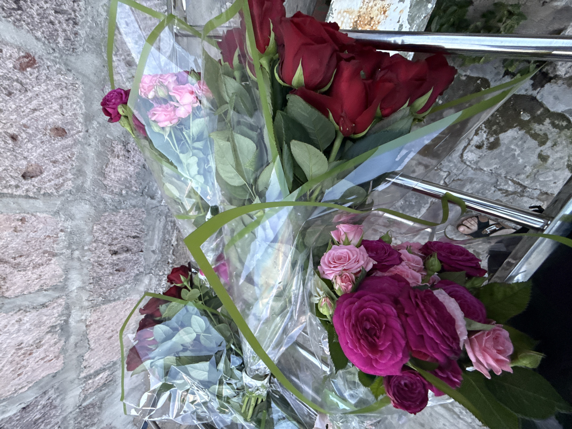 Three bouquets of roses are arranged on stone steps, with a rough stone wall forming the background. One bouquet, on the left, consists of dark red roses. In the center, a bouquet features lighter pink roses. To the right, another bouquet is composed of vibrant red roses. The roses are wrapped in clear plastic with green trim. The steps appear wet in some areas. A pair of sandals is visible on the steps in the lower right portion of the image. The overall scene suggests an outdoor setting in a city. The location in Kosh, Armenia, is not explicitly discernible from the visual information provided in the image alone.