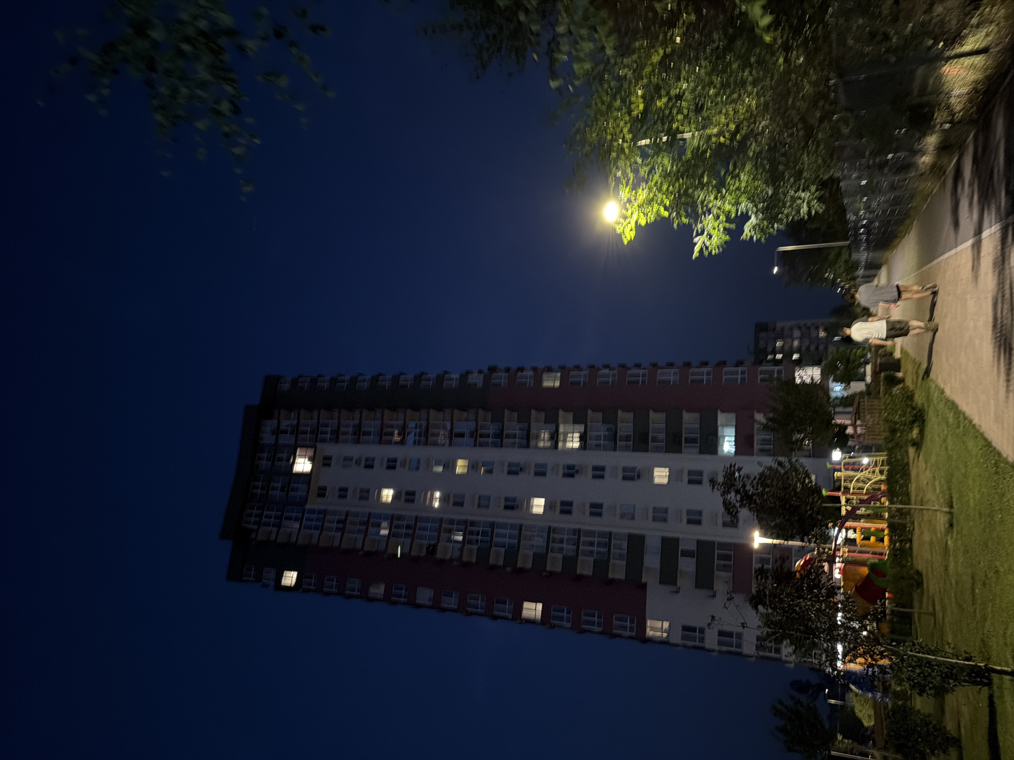 The image depicts a modern apartment building in Kyiv, Ukraine, at dusk. The building is tall and multi-story, with visible lights on in some of the windows. In the foreground, a paved walkway runs alongside a grassy area with trees and a children's playground. Two individuals are walking away from the camera on the walkway. A street lamp illuminates the path, casting shadows from the surrounding trees. The sky is a deep blue, indicating the transition from day to night.
