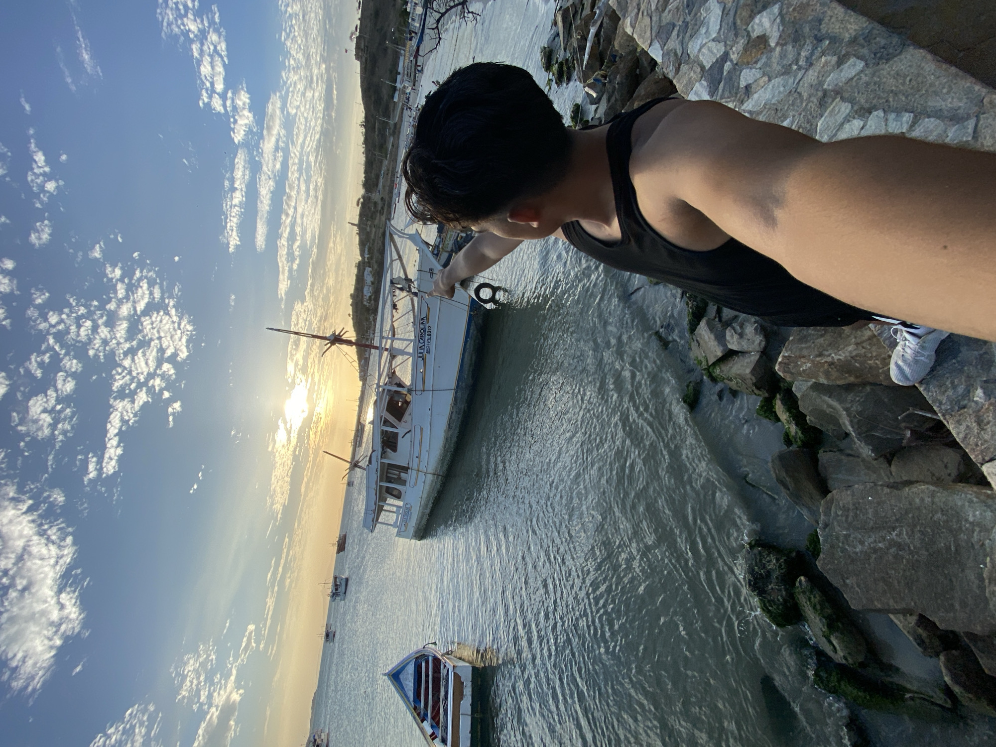 A man wearing a black tank top and white sneakers stands on a rocky bank, pointing towards a white boat labeled "JULIA CAROLINA". The location appears to be near a body of water with other boats in the distance. The sky is visible in the background. This photograph was taken in Juan Griego, Venezuela.