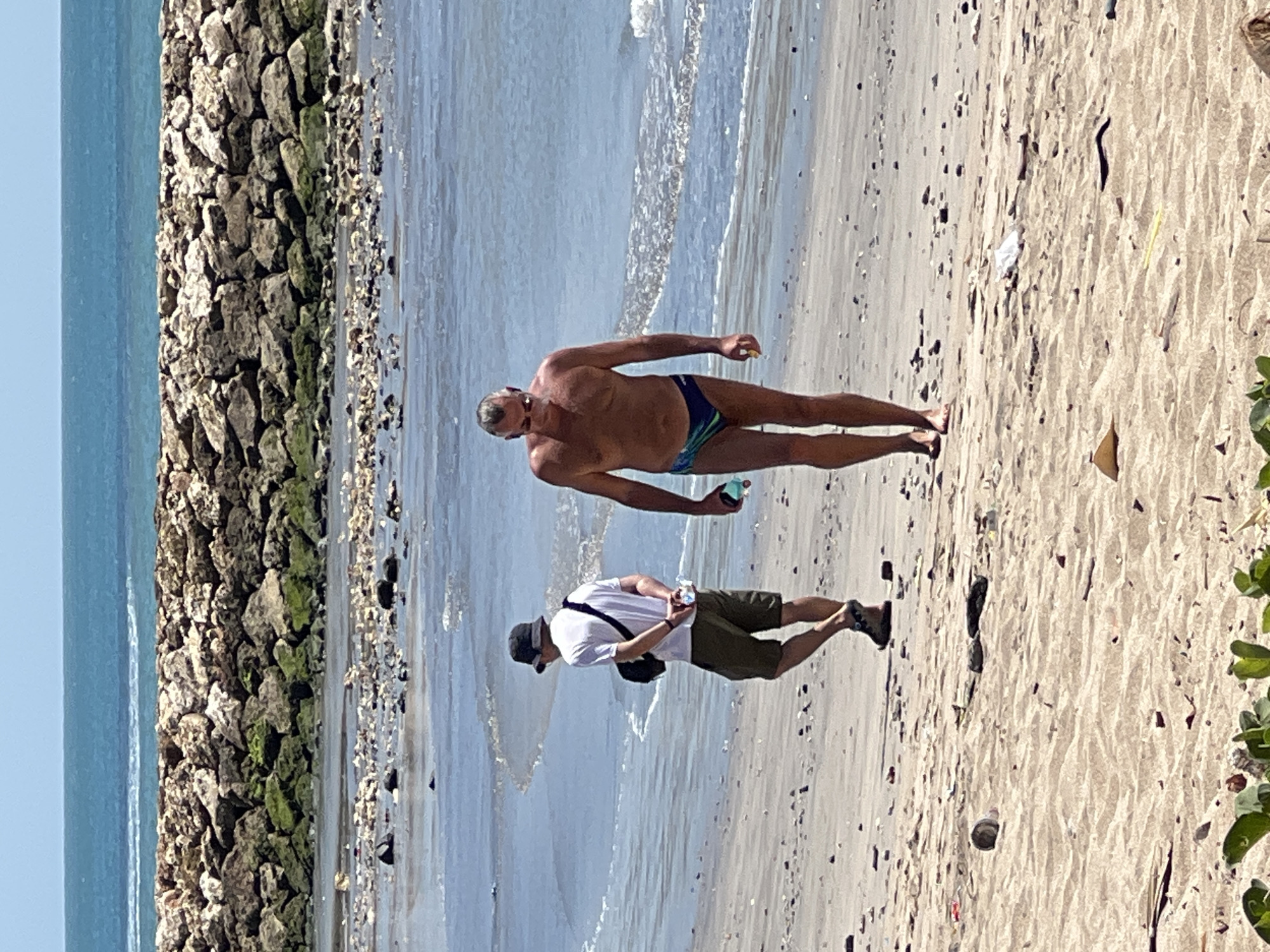 In Kuta, Indonesia, two individuals are observed walking on a sandy beach with the ocean in the background. In the foreground, a man wearing a patterned Speedo and sunglasses is walking towards the right of the frame. To his left and slightly behind him, a person in a white t-shirt, green shorts, and a black hat is walking towards the left. The beach is littered with small stones, debris, and some vegetation in the lower left corner. A rock wall structure extends across the middle ground, separating the beach from the turquoise ocean, which has gentle waves. The sky is clear and blue.