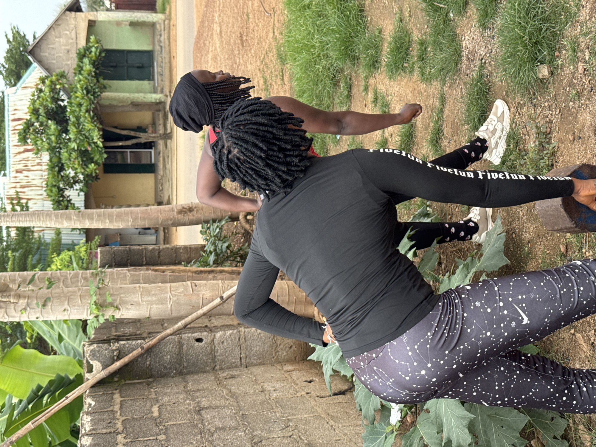 Two individuals are outdoors in what appears to be a residential area in Akwanga, Nigeria. In the foreground, a person wearing a black long-sleeved shirt with white constellation patterns on black leggings and white athletic shoes is bending over. Their back is to the camera, and their arms are extended downwards. Another person, wearing a black beanie and a black long-sleeved top, is standing to the right and behind the first individual, also bending over. The background shows a weathered concrete wall, a dirt path, green grass, and a building with a corrugated metal roof and green and yellow walls. Banana plants and trees are visible in the surrounding environment.
