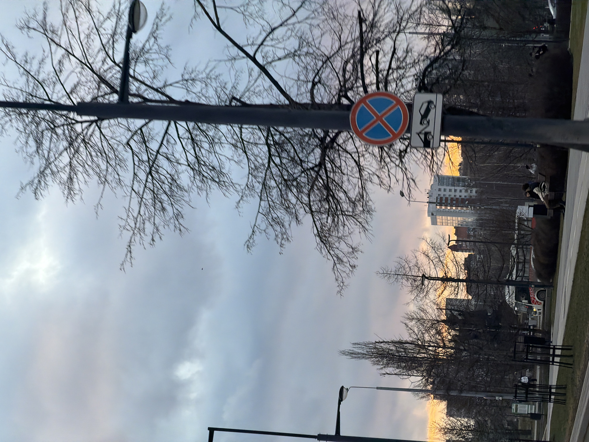 The image depicts a street scene in Perm, Russia. In the foreground, there is a sign indicating no stopping allowed, with an additional sign showing a vehicle being towed. A lamppost runs vertically through the center. Behind the sign, bare trees are visible, silhouetted against a cloudy sky. In the middle distance, buildings are partially obscured by the trees. On the right, a road and some pedestrians are seen, though details are unclear. A bench, possibly made of metal or dark wood, is also visible, with two figures on it. The lighting indicates either sunrise or sunset, with a warm glow in the sky. The orientation of the image is horizontal, and it appears to have been taken from inside a building, looking out through a window.