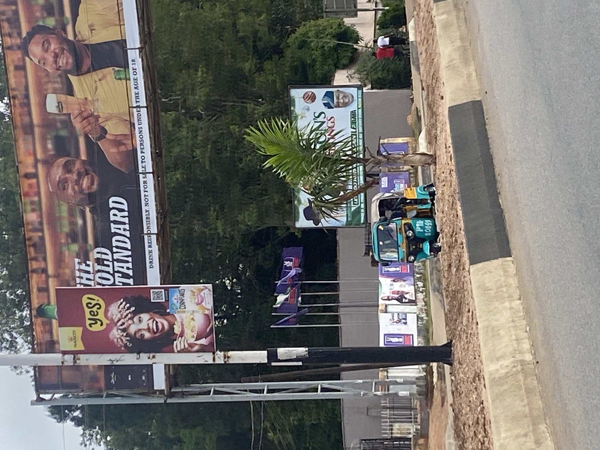 This image captures a vibrant street scene in Jos, Nigeria, during the daytime, likely under an overcast sky or in the late afternoon. The main focus is on multiple billboards displaying various advertisements alongside a roadside.
In the foreground, a large billboard features two smiling Black men. The man at the top wears a yellow top and a black face mask, while the man below him wears a black t-shirt. The man at the top holds a glass with a foamy drink. Prominent text on this billboard reads "THE OLD STANDARD" and a disclaimer: "DRINK RESPONSIBLY NOT FOR SALE TO PERSONS UNDER THE AGE OF 18."
Below this large billboard, a smaller, vertically oriented billboard advertises "Yes! NASCO CORNFLAKES," showing a smiling Black woman with an animated expression, hands near her face, and a bowl of cornflakes.
Further down the road, another large billboard is visible, partially obscured by a palm tree. It appears to show a stylized face in a hat and other graphics, with partial text including "ON'S," "GS," "DOMURA," and "RASAN MA."
On the right side of the road, a blue and white motorized rickshaw, commonly known as a Keke NAPEP in Nigeria, is either parked or in motion. The license plate or identification number "PTA 10149" is visible on its front. The road features a concrete curb separating it from a dirt and gravel shoulder. In the background, green trees and foliage are abundant, suggesting a lush environment. Two distant figures in red and white shirts are visible walking by the side of the road.