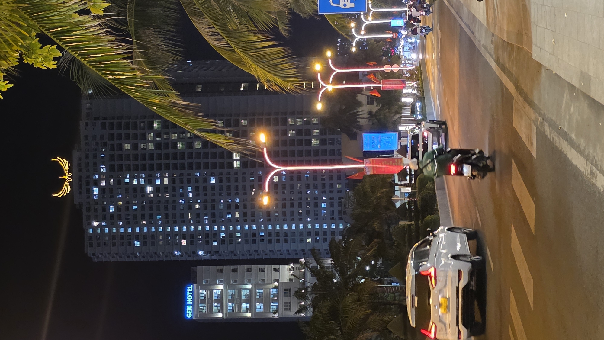 This night-time image captures a bustling urban street scene, likely in Nha Trang, Vietnam, at a slight diagonal angle. On the left side of the frame, a towering, modern building dominates, its facade punctuated by numerous illuminated windows that glow against the dark sky. High on this building, a distinctive golden, stylized logo resembling wings is prominently displayed. Towards the bottom of the frame, on a lower section of the building complex, a bright blue neon sign clearly spells out "GEM HOTEL."
The right side of the image features a wide street with multiple lanes. Several vehicles are visible, including a white SUV in the foreground and a person riding a motorcycle or scooter, wearing a green top, further down the road. Another motorcycle's taillights can be seen in the distance. The street is brightly lit by decorative streetlights, which feature unique Y-shaped structures with glowing orange spherical lights and linear red light elements. Palm tree fronds are visible at the top and bottom edges of the frame, indicating a tropical or coastal setting. A blue road sign with an unidentifiable white symbol is also present on the right. The overall scene depicts a lively and well-lit city environment after dark.