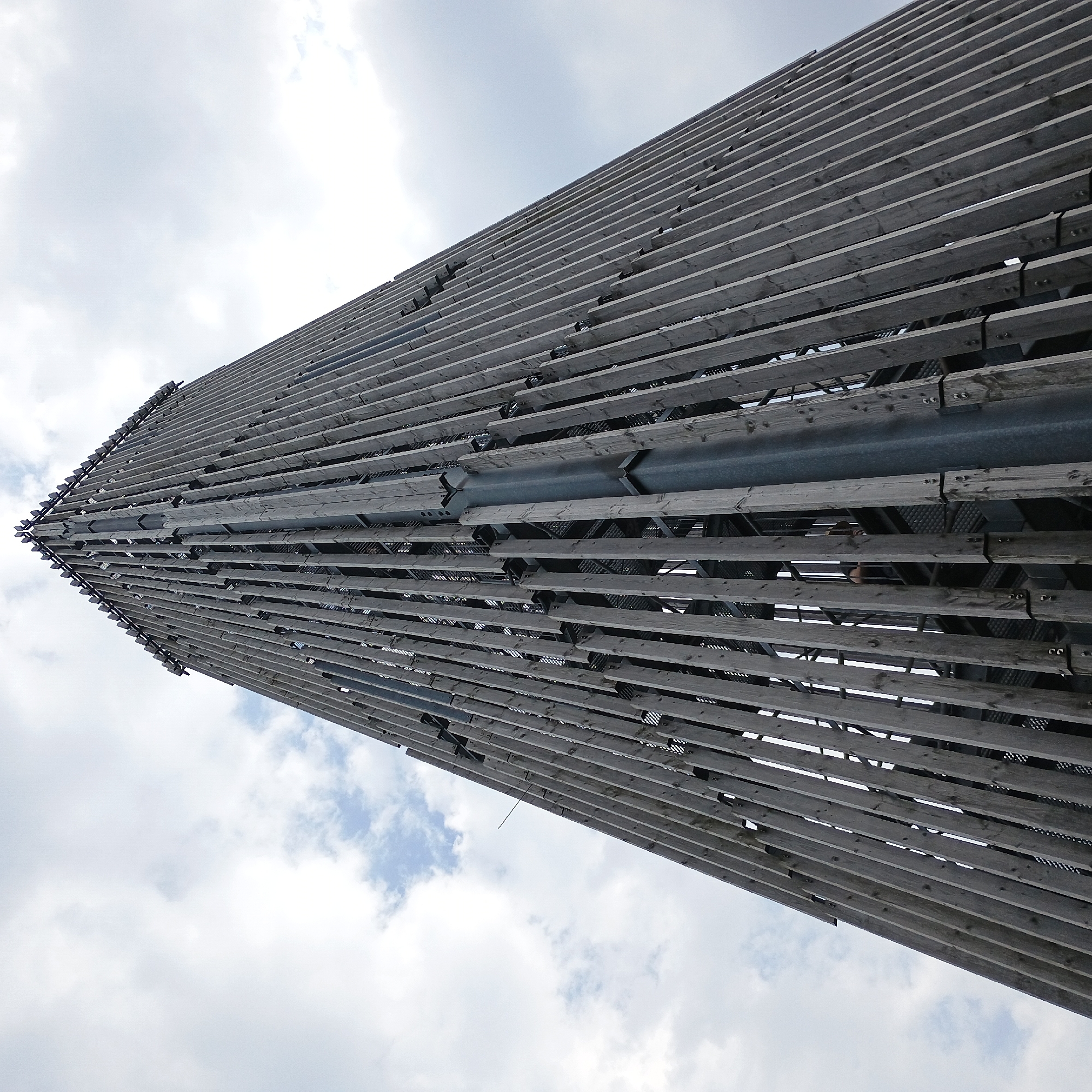 This is a low-angle, full-frame shot of the Möhnesee viewing tower, a tall, tapering structure predominantly made of weathered wooden planks and a central metal column. The tower is set against a cloudy, overcast sky. The wooden planks are vertically oriented and run the height of the structure, with metal supports visible between them. A spiral staircase is discernible within the open framework of the tower. In the mid-section of the tower, a person, seen from the back, is ascending the stairs. The tower's construction suggests a modern design with an emphasis on natural materials, likely located in the vicinity of the Möhnesee reservoir in Germany.