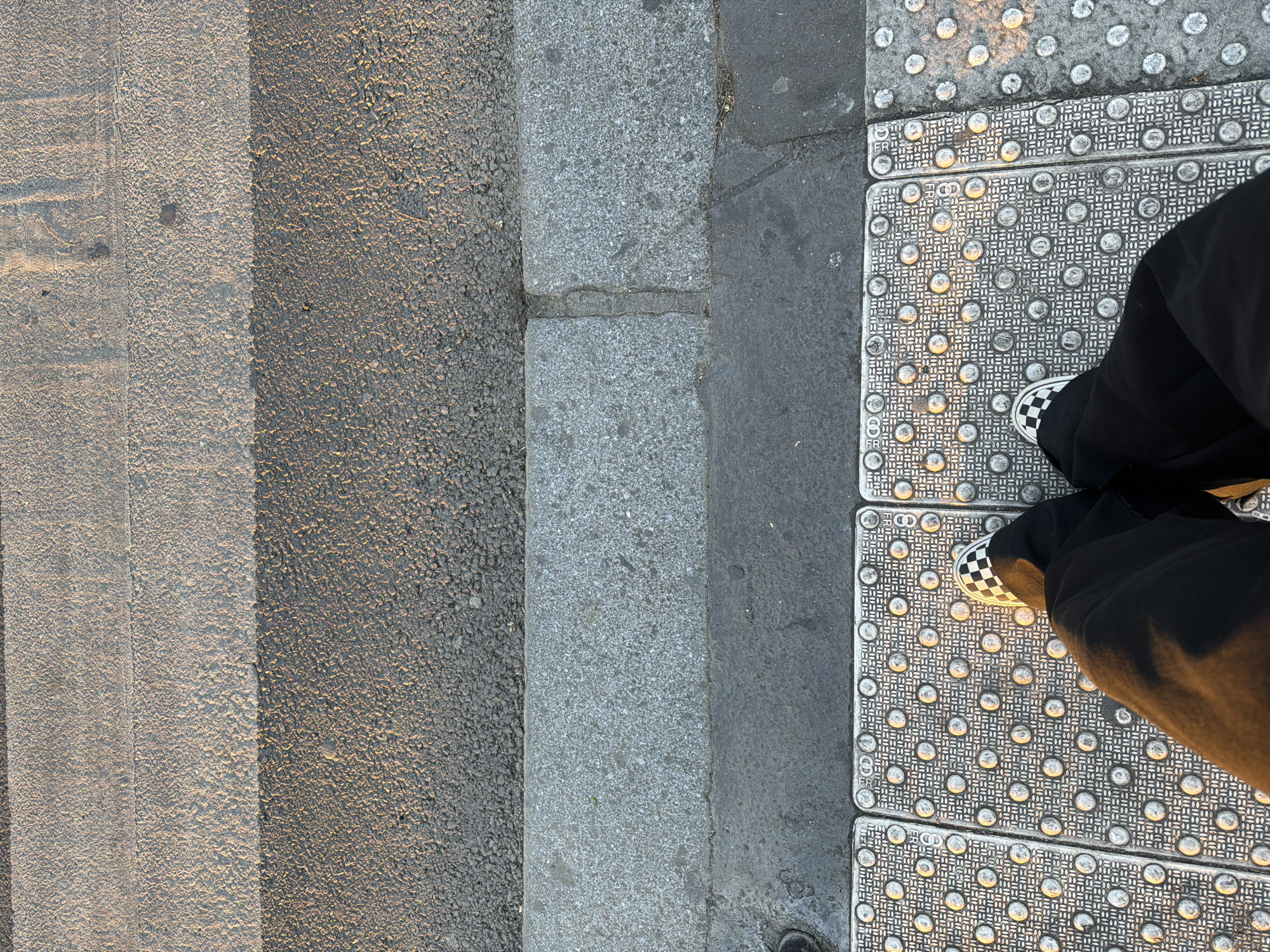 This high-angle, downward shot captures a pedestrian's feet on a sidewalk, next to a street, in what is identified as Paris, France. The scene is illuminated by warm, directional sunlight, suggesting either early morning or late afternoon, creating distinct shadows across the ground.
The image is divided into several distinct textures and surfaces. On the far left, a section of an asphalt road with faint, light-colored horizontal markings (possibly part of a crosswalk) is visible. A grey concrete or stone curb forms the boundary between the road and the sidewalk. The sidewalk itself features prominent metal or concrete tactile paving, characterized by a grid of raised, truncated domes designed to aid visually impaired pedestrians.
A person is standing on this tactile paving, with their lower legs and feet visible. They are wearing black trousers and distinctive black and white checkered slip-on shoes, resembling Vans. One foot is positioned towards the bottom right corner of the frame.
The sunlight highlights the raised domes of the tactile paving, making them appear brighter, while the grooves and the areas under the domes are in shadow. The curb and parts of the street closer to the sidewalk also show variations in light and shadow. The letters "FR" are visibly embossed into the material of the tactile paving along its edge, serving as a clear location cue for France. The overall composition offers an intimate glimpse of street-level urban detail and pedestrian interaction with city infrastructure.