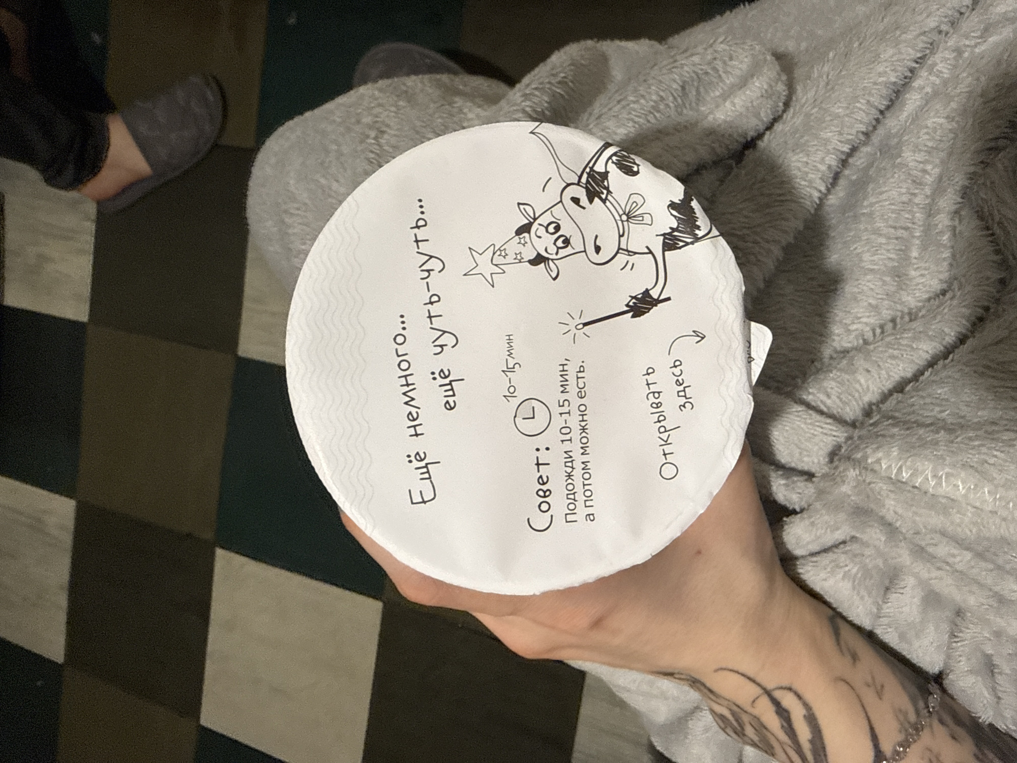 A hand is holding a round, white container with Russian text and a cartoon drawing of a cow wearing a wizard hat and holding a wand. The text says "Еще немного... еще чуть-чуть..." (A little more... a little bit more...), with a suggestion to wait 10-15 minutes before eating, and instructions to open it at the indicated spot. The hand has a tattoo on the wrist and lower arm. The person holding the container is sitting or reclining, as evidenced by the grey, plush material in the background. A checkered floor, and a foot wearing a gray sock are also visible. There's no direct information to place the scene in Moscow, Russia.