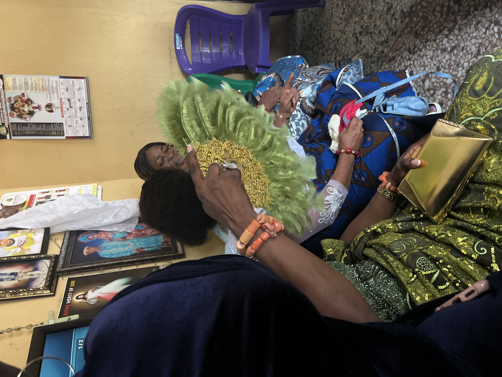 The image captures a scene in what appears to be a room in Warri, Nigeria. Several people are visible, with a focus on two individuals holding a green feathered fan with gold accents. One person is wearing a blue shirt, and another is wearing a green and gold patterned fabric. A woman with dark hair is smiling in the background.
There are items scattered around, including blue and brown patterned fabric, a blue purse, a gold clutch, and a measuring tape. In the background, a purple plastic chair is visible.
On the wall, there are religious pictures framed along with what appears to be a calendar, and a framed poster. A television screen shows "1/2" in the lower left corner. The floor has a patterned tile surface.