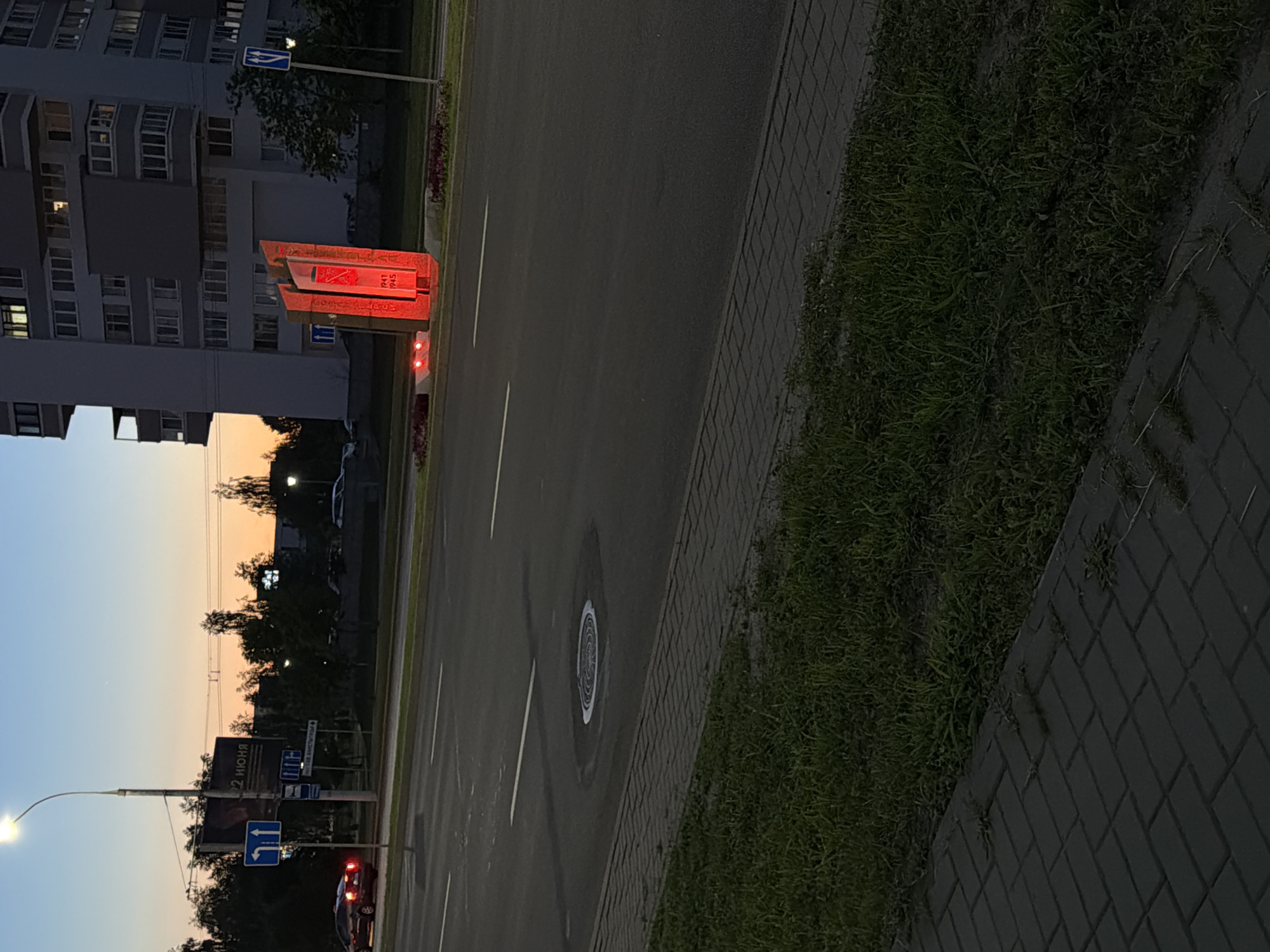The image shows a monument illuminated in red, located on a street in Brest, Belarus. The monument appears to be made of metal and has the years "1941" and "1945" inscribed on it. It is situated in front of a modern apartment building. To the left of the monument, a street with traffic lanes is visible, with a car driving on it. There are also street signs, a lamppost, and trees lining the street. The sky is a dusky blue and orange, suggesting it is either early morning or late evening. The ground in the foreground is paved with gray interlocking bricks, adjacent to a strip of green grass. The overall scene depicts an urban environment with a memorial.