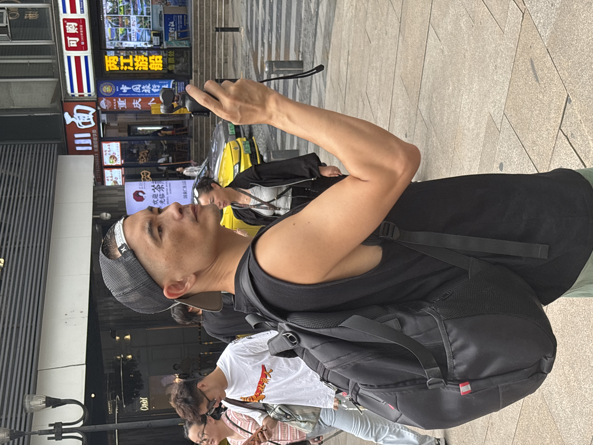 A man with a gray cap and black tank top is holding a camera and taking a picture on the street in the Yuzhong District of Chongqing, China. He is wearing a large black backpack. Behind him are several people walking, including a woman in a white shirt with a graphic print, and a person in a black jacket over a white shirt. To the left, buildings with Chinese-character signage are visible. Across the street, a yellow taxi is parked. The environment is a paved public walkway.