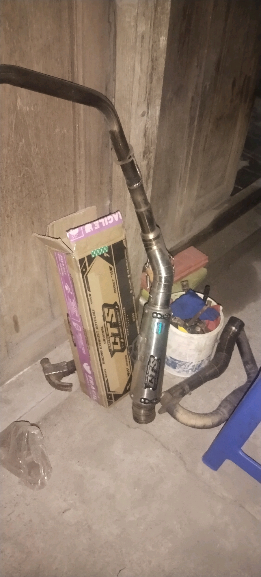 The image shows an exhaust pipe and muffler, along with a cardboard box, a bucket, and other miscellaneous items, arranged on a concrete floor against a wooden wall. The exhaust pipe is made of metal and appears to be curved. The muffler is cylindrical and also made of metal, with some branding visible. The cardboard box is open and has some text and graphics printed on it. The bucket contains some blue and red fabric items. A blue plastic object, possibly a stool or chair, is partially visible on the right side of the image. The overall environment appears to be an outdoor or semi-outdoor space, possibly a garage or workshop, given the presence of tools and construction materials. The lighting suggests it is either nighttime or a dimly lit area.