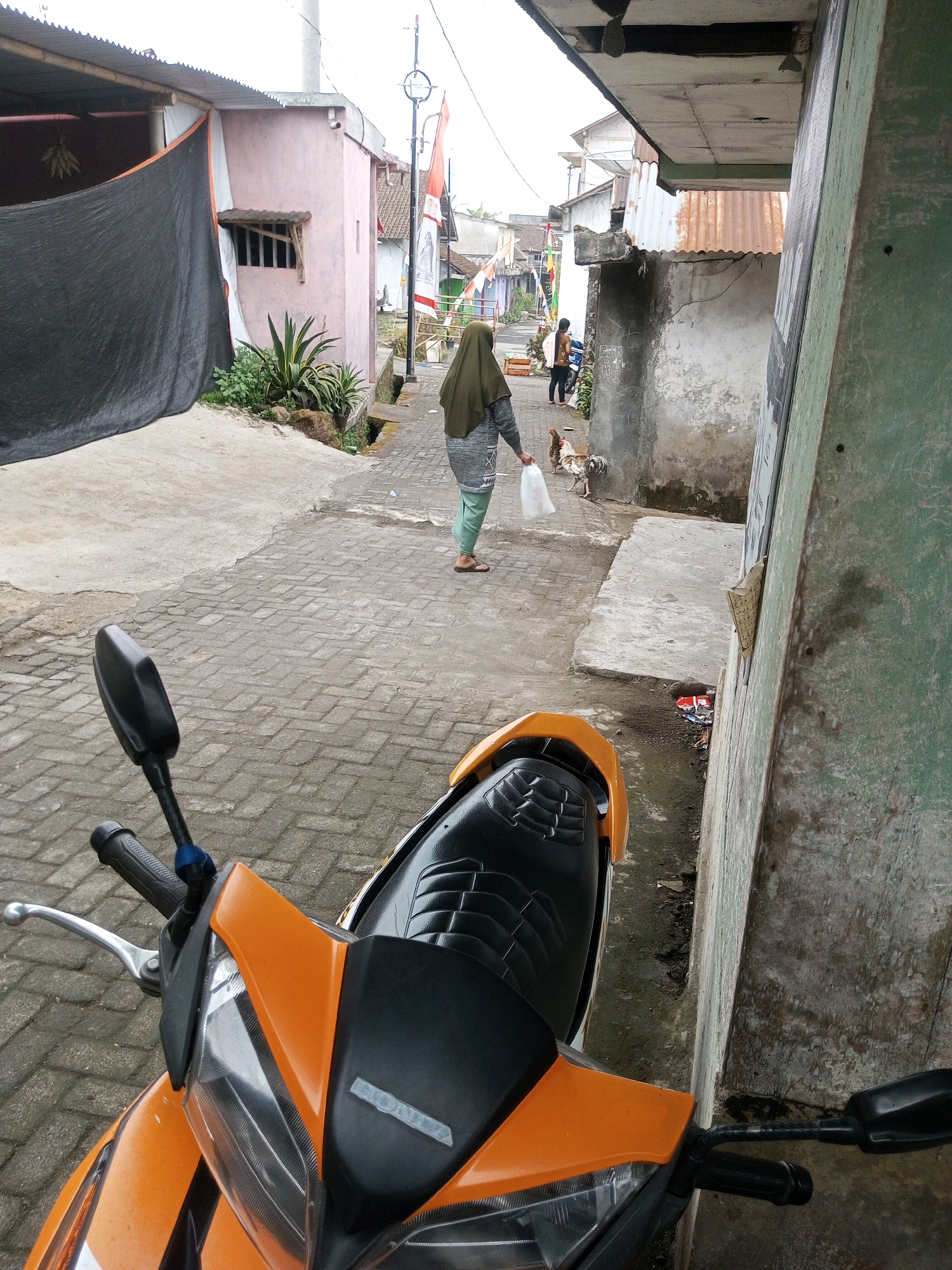 A woman wearing a green hijab and a grey patterned shirt walks down a paved alley in Andongsili, Indonesia. She carries a white plastic bag in her right hand. A rooster stands to her left, and another person is visible further down the alley in the background. The alley is lined with concrete buildings, some with tin roofs, and vegetation grows along the sides. A motorcycle is parked in the foreground, partially obscuring the view.