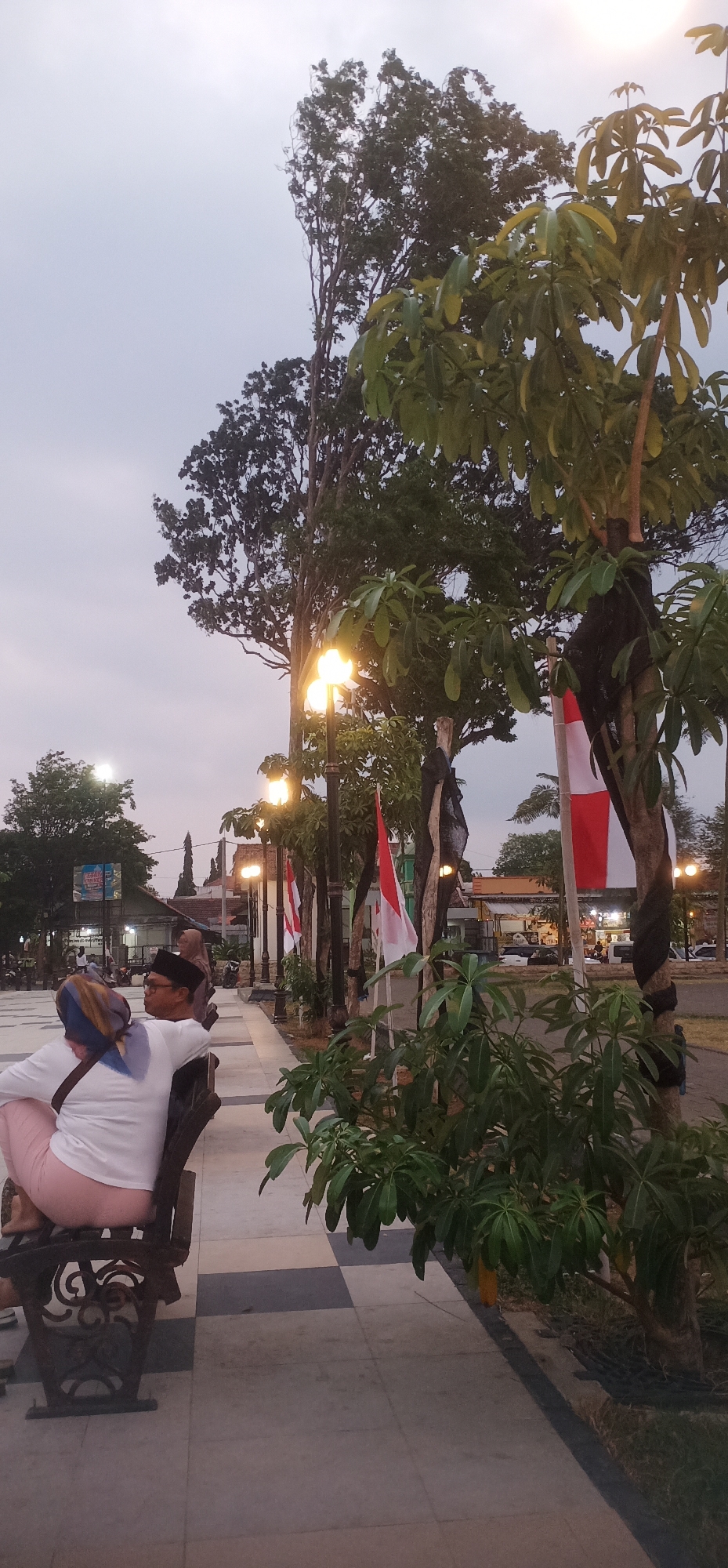 A medium shot shows an outdoor scene in the city of Probolinggo, Indonesia, at dusk. The foreground features a bench with two people sitting on it, facing away from the camera. The person on the left wears a white long-sleeved shirt and a blue and tan headscarf. The person on the right wears a white long-sleeved shirt and a black peci hat and glasses. Behind them, a checkered tiled walkway leads to a street lined with trees and lampposts. Indonesian flags, red and white, are tied to some of the trees. In the background, buildings and shops are visible, with cars and people in the distance. The sky is overcast and a pale grey. The scene appears to be a public space, possibly a park or plaza, illuminated by the warm glow of the lampposts.