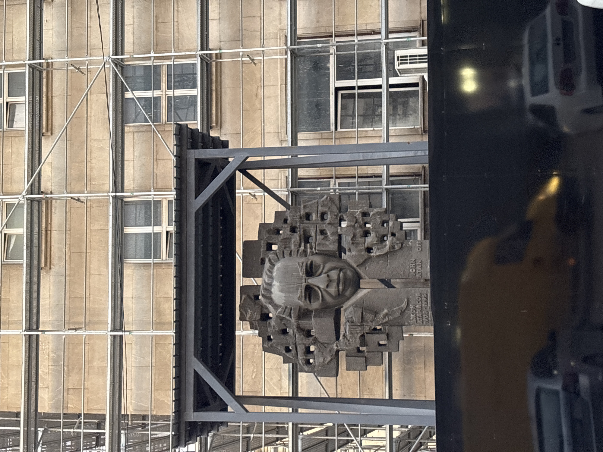 The image shows a bust sculpture of a man wearing glasses, positioned under a metal canopy. The sculpture is made of dark material and features intricate detailing around the head, resembling blocks or abstract shapes. Below the bust, text is visible, including "ATANASOFF 1903-1995" and "JOHN ATAN OFF". The sculpture is situated in front of a building with a stone facade and multiple windows, which is partially covered by scaffolding. In the foreground, there is a dark, reflective surface showing the blurred reflections of vehicles, including a white car. The overall scene suggests an outdoor public space in Sofia, Bulgaria, likely a monument or memorial.