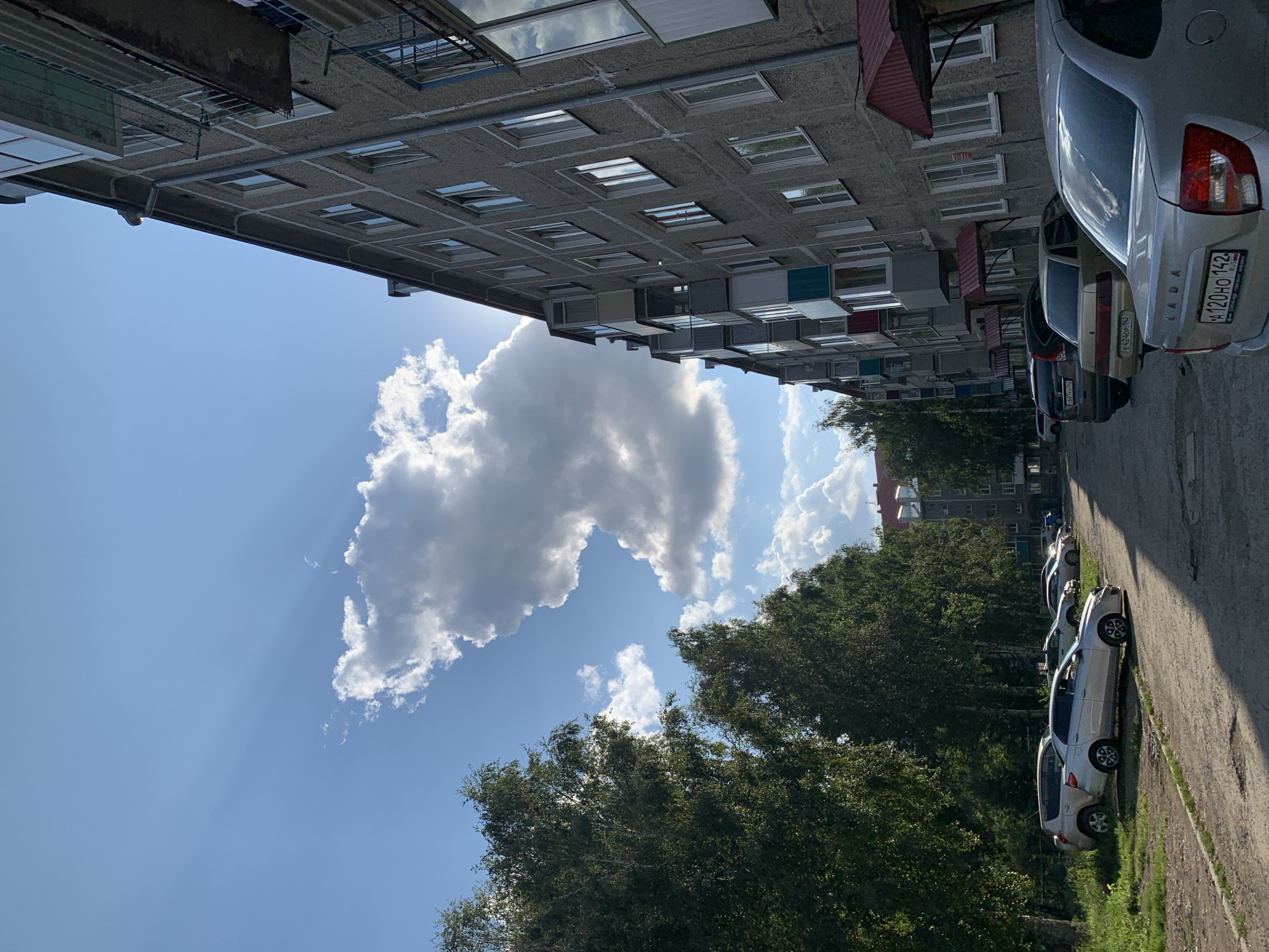 The image depicts a typical urban scene in Prokopyevsk, Russia, likely on a sunny day with some clouds. The foreground is dominated by a row of parked cars along a paved area, with a gray, multi-story apartment building to the right. The building features numerous windows and balconies, some with colorful coverings. To the left, a line of green trees with dense foliage lines the edge of the paved area and extends into the background. Sunlight streams through a gap in the clouds, casting shadows and highlighting parts of the building and trees. A few other buildings are visible in the distance, including what appears to be a red-roofed structure. The sky is blue with several white and gray clouds, and sunbeams are visible piercing through the clouds. The overall impression is a snapshot of everyday life in a residential area of the city.