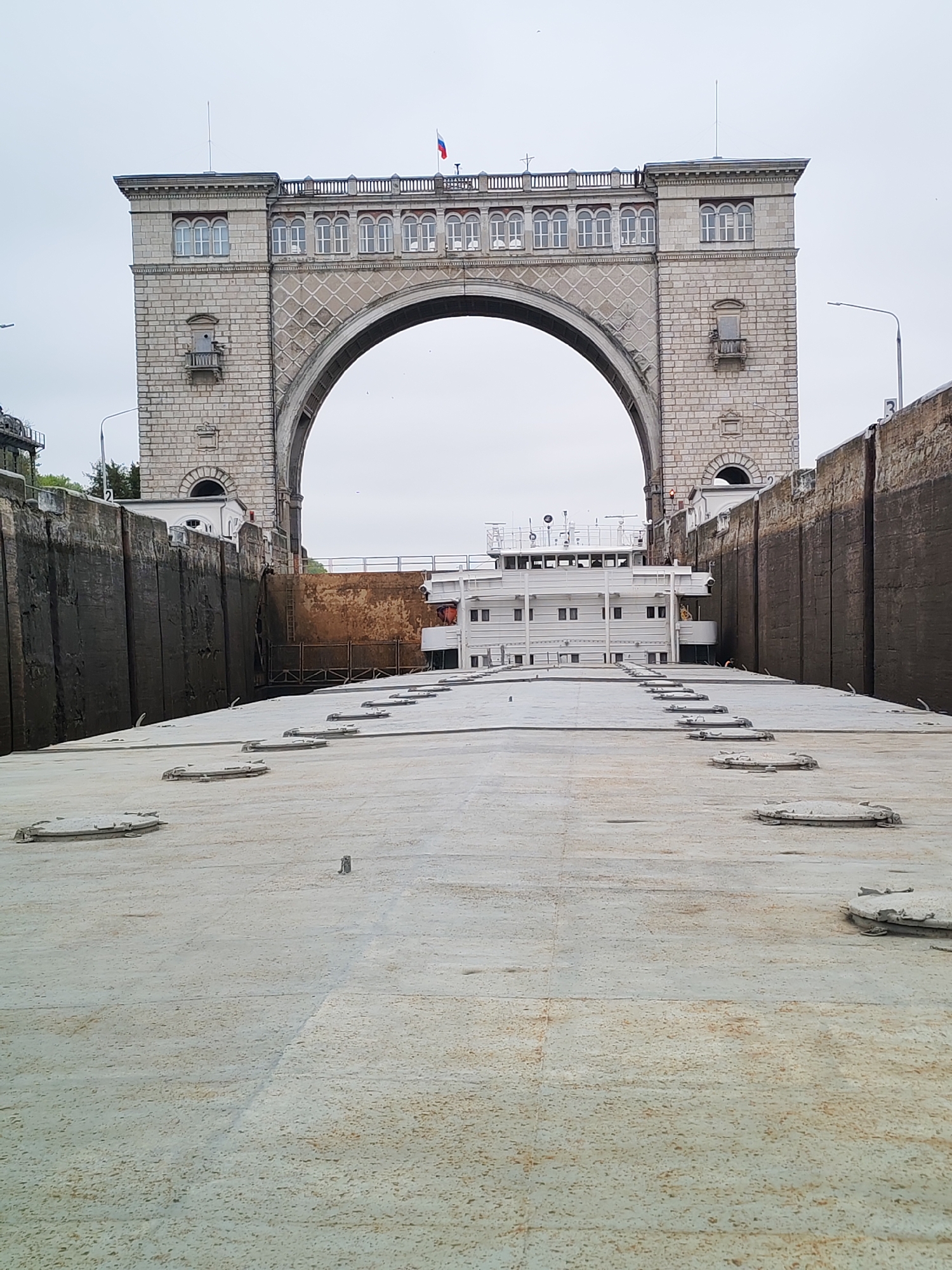 The image depicts a boat navigating a lock in an urban area, possibly a canal or river system. In the background is a large, arched stone structure with architectural details including windows, balconies, and a colonnaded top. A flag, possibly Russian, is visible atop the arch. The lock walls are dark and appear damp. The boat is centered within the lock, with its flat top covered in numerous circular access hatches. The sky is overcast. The location is in the city of Unknown, Unknown.