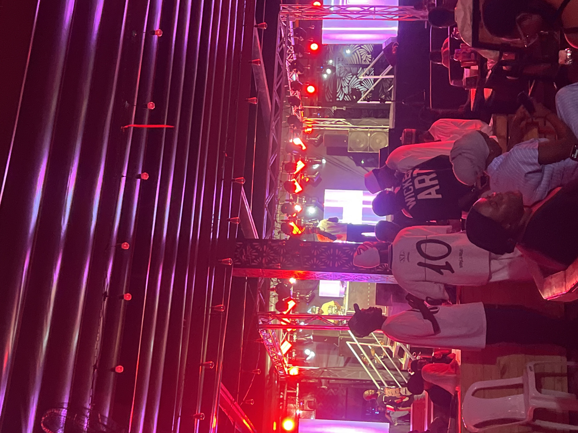A vertically oriented photograph depicts a scene on a stage in what appears to be a club or concert setting, potentially in Howayi, Nigeria, given the prompt's location mention. The lighting is dominated by red and purple hues. A metal structure composed of vertical bars and small round red lights is visible on the left side of the image. Overhead, metal scaffolding supports stage lighting and equipment.
A group of people is gathered on and around the stage. One individual is wearing a white shirt with a prominent number "10" printed on the back. Another wears a black shirt with the partial word "WICKED" and the letters "ART" visible. There are other individuals partially obscured or with their backs to the viewer.
Some people appear to be seated on chairs, some of which are white plastic. The general impression is of a crowded, energetic atmosphere, possibly during a performance or concert. The scene is backlit, making details somewhat difficult to discern.