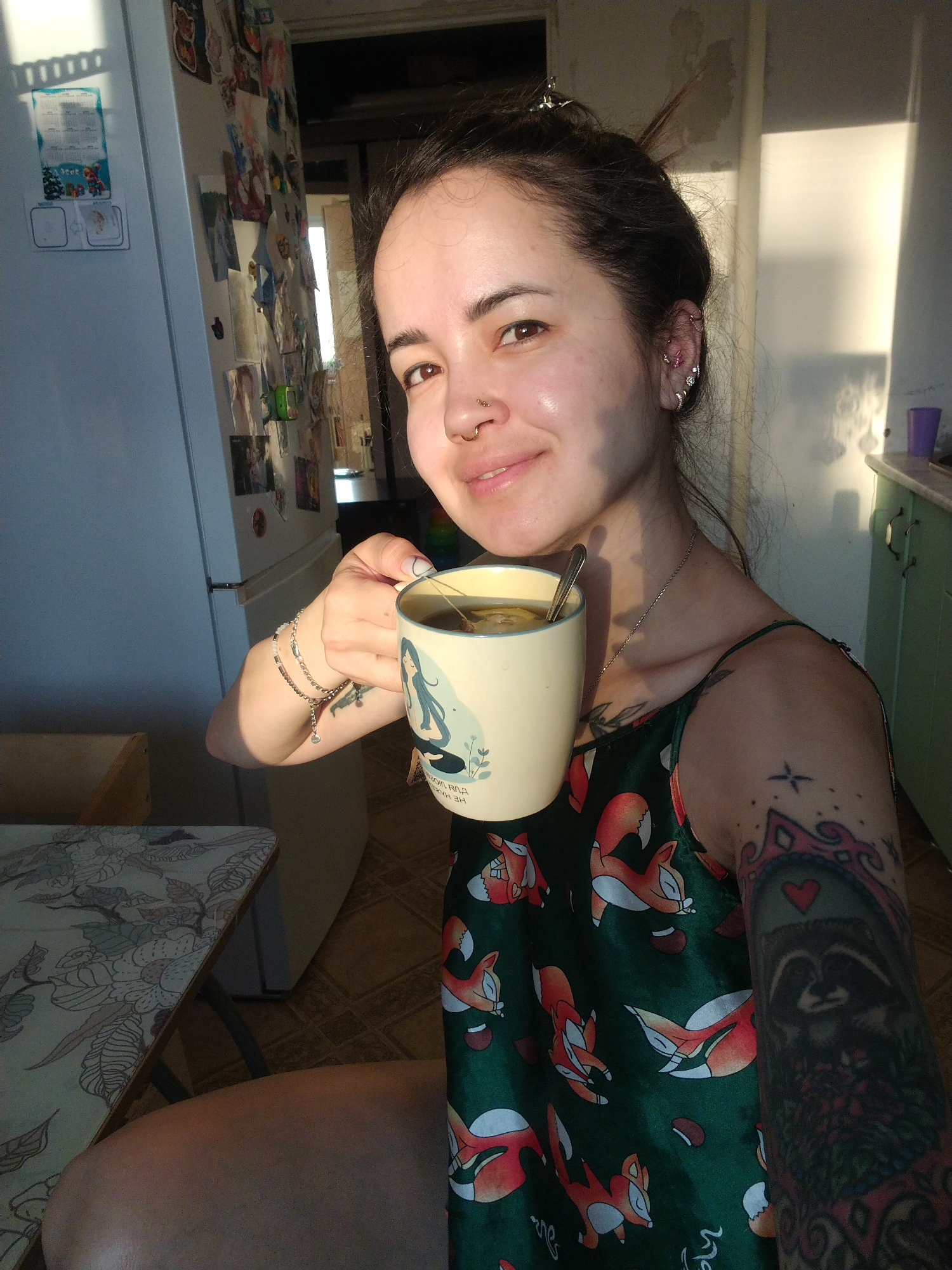 A woman is posing for a photo while holding a mug. She is fair-skinned with dark hair pulled back, and has piercings in her ear and nose. She wears a thin chain around her neck and several bracelets on her left wrist. A tattoo is visible on her left arm. She wears a dark green top with a pattern of orange foxes. The mug she holds is light yellow and features a drawing of a woman. There is a spoon in the mug.
Behind her is a kitchen area. A refrigerator with papers and images stuck to it is visible. Next to the refrigerator, a partially open doorway reveals a sunlit area beyond. A cabinet is visible to the right of the woman. The scene is indoors and appears to be lit by natural light. The location is Noviy, Russia.