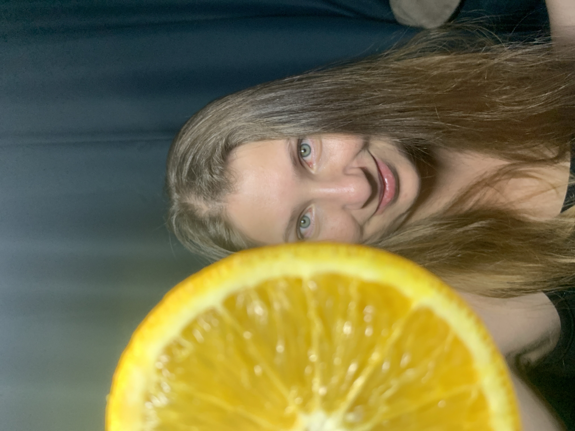 A woman with light brown hair and blue eyes is holding half of an orange in front of her face. The orange is in focus, and the woman is smiling slightly. The background is a dark blue curtain. The location in the city of Chaikovsky, Russia is not determinable from the image.