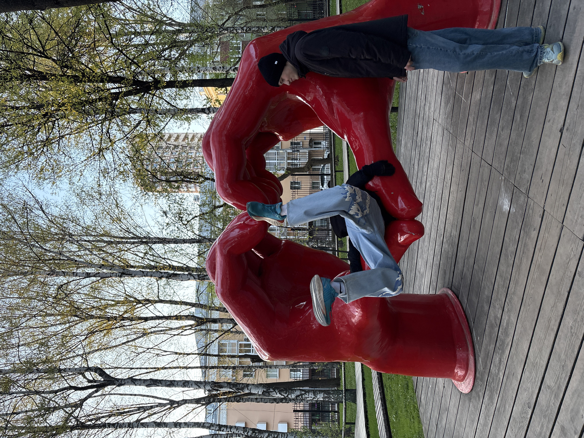 Two individuals are interacting with a large, red sculpture of two hands intertwined in Perm, Russia. One person, wearing a black jacket and beanie, is resting their upper body on the upper hand. The other person, wearing light blue jeans and blue sneakers, is positioned in the lower hand of the sculpture. The sculpture is on a wooden platform. In the background are buildings and trees with budding leaves. The sky is visible through the trees.