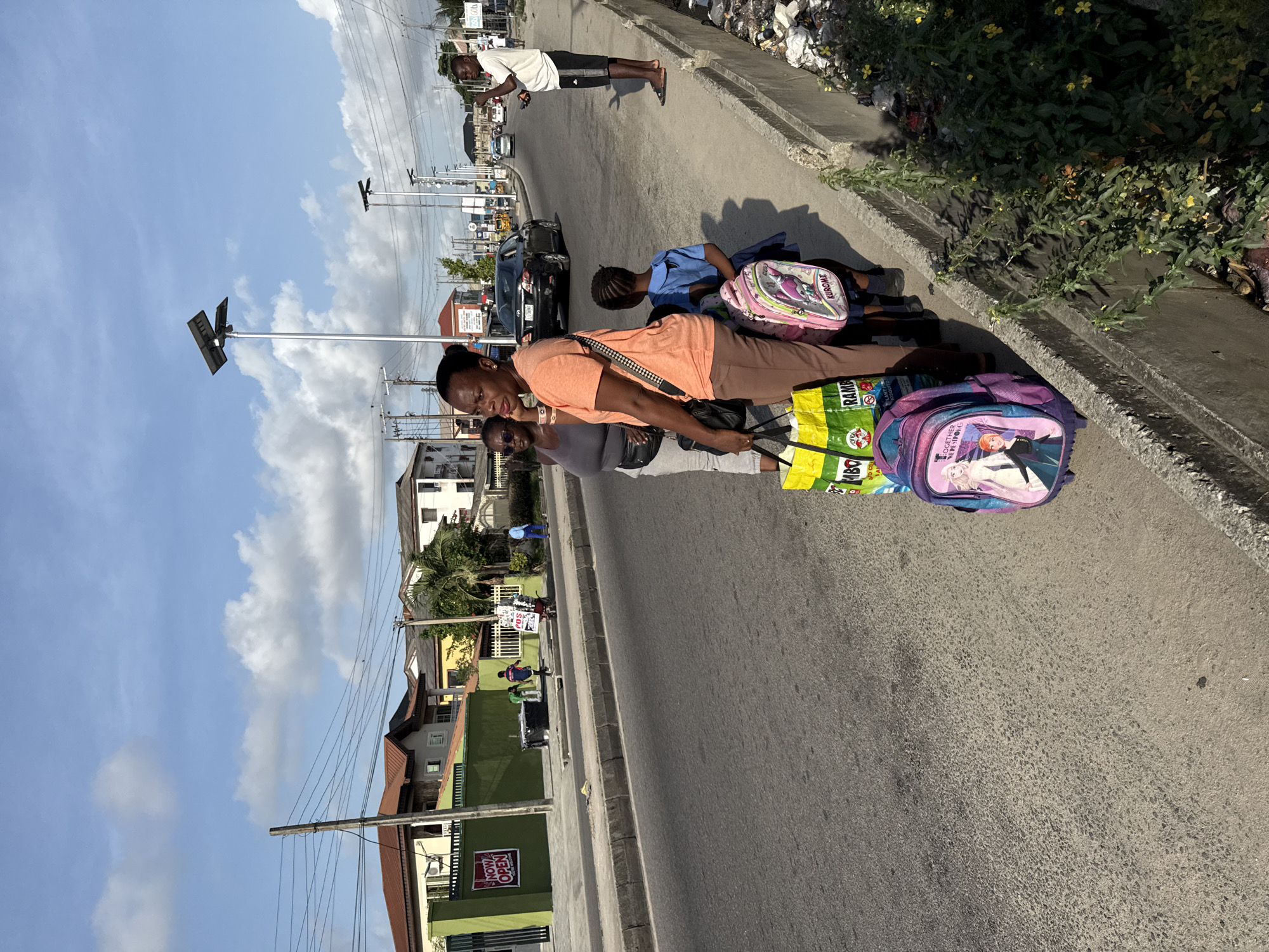 A street scene in Ado, Nigeria. A woman in an orange top and brown pants stands on the roadside with a child in a blue uniform, both leaning on backpacks. Another woman in sunglasses stands nearby. A black car passes on the road, with another person walking in the distance. Buildings and utility poles line the street. The sky is partly cloudy. A concrete curb separates the road from a strip of vegetation and debris.