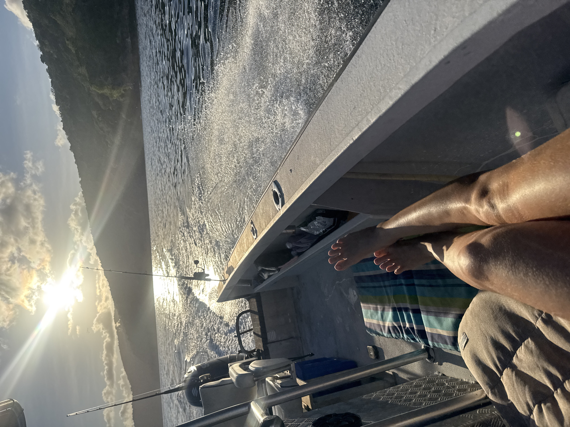 A person's tanned legs and bare feet are visible in the foreground, resting on a striped cushion and a light brown cushion. The person appears to be on a boat, as indicated by the metal railing and seats in the left and background. Two fishing rods are propped up on the boat. The boat is moving through choppy, sun-dappled water, leaving a wake behind it. In the background, a lush, green, mountainous landscape rises from the water. The sun is low in the sky, casting a warm glow and lens flares across the scene. The overall impression is one of a relaxed day spent fishing on the water, possibly in a tropical or coastal area of Australia, given the vegetation and lighting.