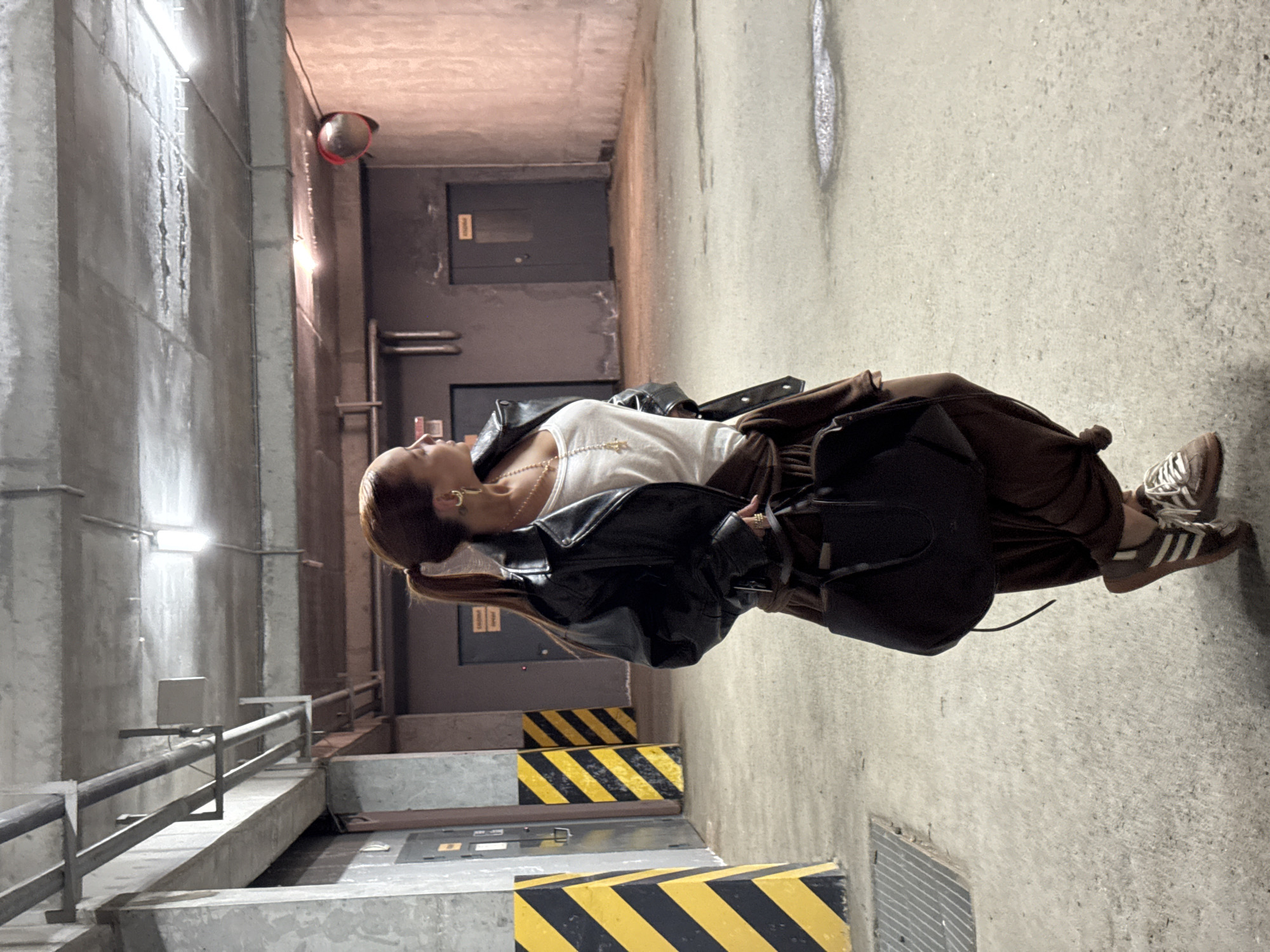 A young woman is posing in an urban setting. She is wearing a white top, brown pants, a black leather jacket, and Adidas sneakers. She also has a black bag. The location appears to be an underground parking garage or service area with concrete walls, railings, and hazard markings. A door and a reflective security dome are visible in the background. It is not possible to verify the city of Izhevsk, Russia, as the location based on the image alone.