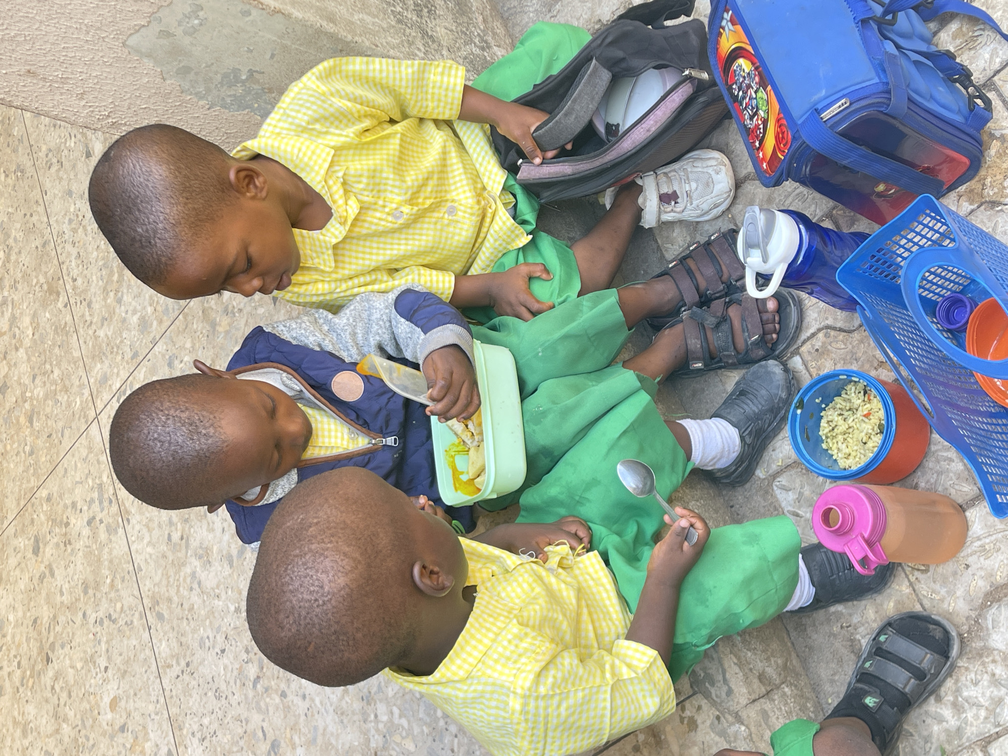 Three young children are sitting on a tiled surface in Akwanga, Nigeria, surrounded by their belongings. Two of the children are wearing yellow checkered shirts and green pants. One child wears a similar outfit with a navy blue jacket layered over the shirt. They appear to be eating lunch, with one child holding a spoon and another examining a container with food. Around them are lunchboxes, water bottles, a blue basket, and shoes.