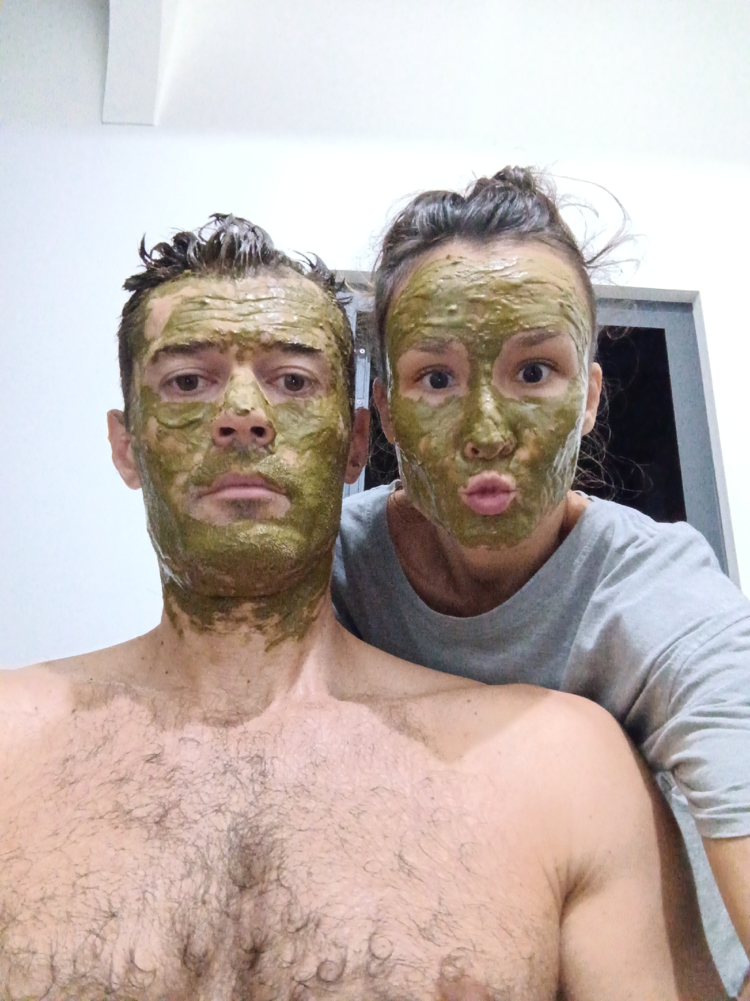 A close-up selfie shows two light-skinned people with green face masks. The person on the left is a shirtless man with short dark hair plastered to his forehead. He has a moderate amount of chest hair. The person on the right is a woman wearing a gray shirt and puckering her lips. Her dark hair is pulled back into a messy bun. They appear to be indoors, with a white wall and a dark doorway visible in the background. The location is unknown.
