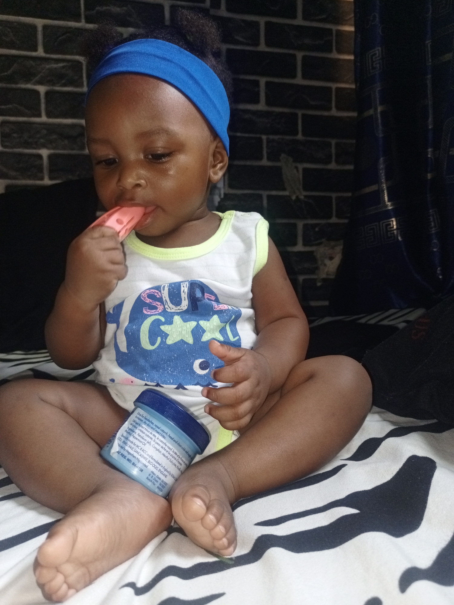 A young child with dark skin, wearing a blue headband and a white and blue graphic t-shirt, sits cross-legged on a patterned bedspread. The child is holding a pink teething toy or similar object and has it in their mouth, looking downwards with a focused expression. Their t-shirt features a design that appears to read "SU C★★", likely part of "SUPER COOL" or similar.
Between the child's legs rests a small, light blue jar with a dark blue lid. The label on the jar is partially visible, showing text in English and French. The English text includes "Use for headaches, nasal catarrh, head and chest colds and irritation, dry skin and insect bites." and "Ingredients: Petroleum Jelly, Camphor, Menthyl Salicylate, Menthol, Peppermint Oil." It also states "Specially made for MC & KCC and formulated in England by SG LTD, IRWELL VALE, LANCASHIRE, BLO OGG, ENGLAND. Distributed by Megachem Nig Limited. AC REG. NO: B4-4138. Man. Date: 06/2012. B. B. Date: 06/2020. Batch No: MX2254."
The setting appears to be indoors, possibly a bedroom. A dark brick-patterned wall is visible in the background on the left, while dark blue curtains with a gold geometric pattern hang on the right. The child is sitting on a white blanket or sheet with black, irregular stripe patterns. A dark, possibly fabric object or bag, is next to the child on the right. The lighting suggests an indoor environment, but the specific time of day is not discernible.