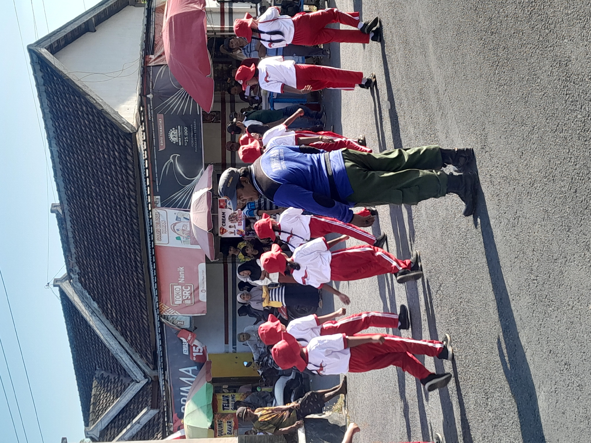 The image depicts a street scene in Bulurejo, Indonesia, where a group of children in matching red hats and white shirts with red accents are marching. They are wearing red pants with white stripes. One adult in a blue jacket and green pants, wearing a dark cap and boots, appears to be leading or supervising the children. Behind them, several adults are visible, some observing the march. Various stalls and awnings line the street, suggesting a marketplace or event. The architecture of the buildings features dark tiled roofs. The ground is paved asphalt, and shadows from the people and overhead structures are cast across it, indicating a sunny day. A motorcycle is parked on the left side of the street, and a man is seated on it.