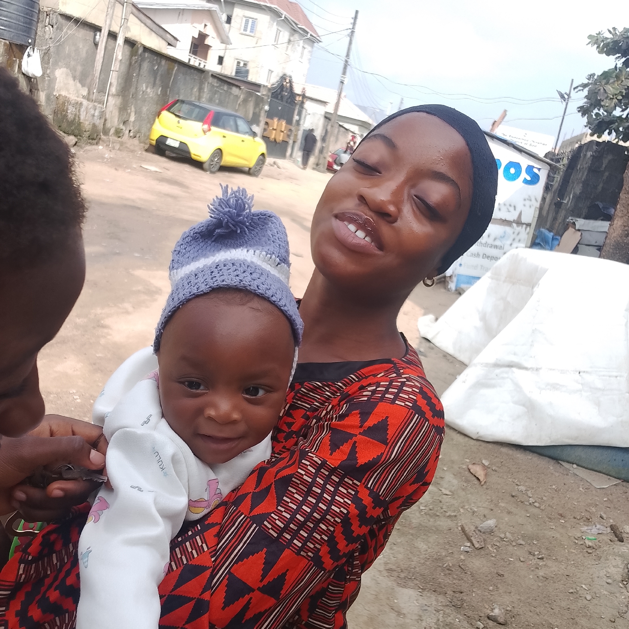 The image shows a young woman holding a baby outdoors in what appears to be an urban environment. The woman is wearing a black headscarf and a red and black patterned top. She is looking upwards with her eyes closed. The baby is wearing a white outfit and a blue and white striped knitted hat with a pom-pom. The baby is facing forward, looking at the camera with a slight smile. The background shows a dusty street with buildings and a yellow car parked in the distance. There are also some large white bags and debris scattered on the ground. The overall impression is of a casual outdoor scene, possibly in a residential area. The image is taken from a slightly high angle, looking down at the subjects. The lighting suggests it is daytime. The woman appears to be in her late teens or early twenties, and the baby is an infant. Their interaction seems affectionate as the woman holds the baby close. The location is stated to be Ado, Nigeria. The environment suggests a mix of residential and commercial activity, with unpaved ground and modest structures.