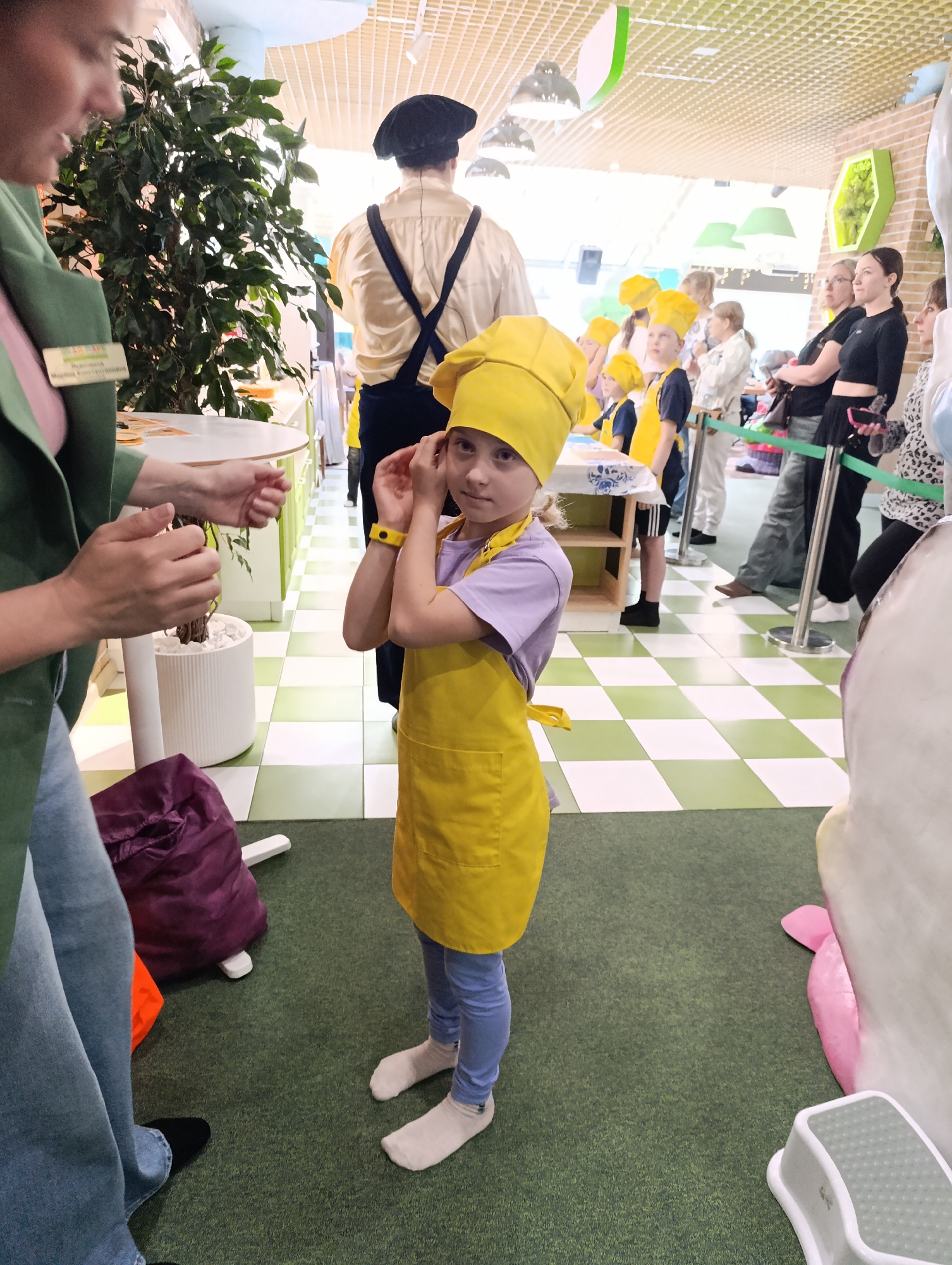 A full shot captures a young girl standing indoors. She wears a yellow chef's hat and apron over a light purple shirt and blue leggings, with white socks on her feet. She holds her hands near her face, appearing to adjust the hat, and wears a yellow wristband.
To her left, a woman is partially visible, wearing a green blazer and jeans. A ficus tree in a white planter and a plum-colored bag are also nearby. The flooring under the girl is a carpet.
Behind the girl, in the center of the image, is a man in a vintage costume with suspenders and a black hat. Further back, a line of children in yellow chef's hats and aprons is visible, along with several adult women standing in line.
The floor in the background is a light green and white checkerboard pattern, and the ceiling has a grid-like structure with pendant lights hanging down. A green honeycomb-shaped decoration is attached to the wall in the background. To the right of the girl, a large, white, abstract object is visible.