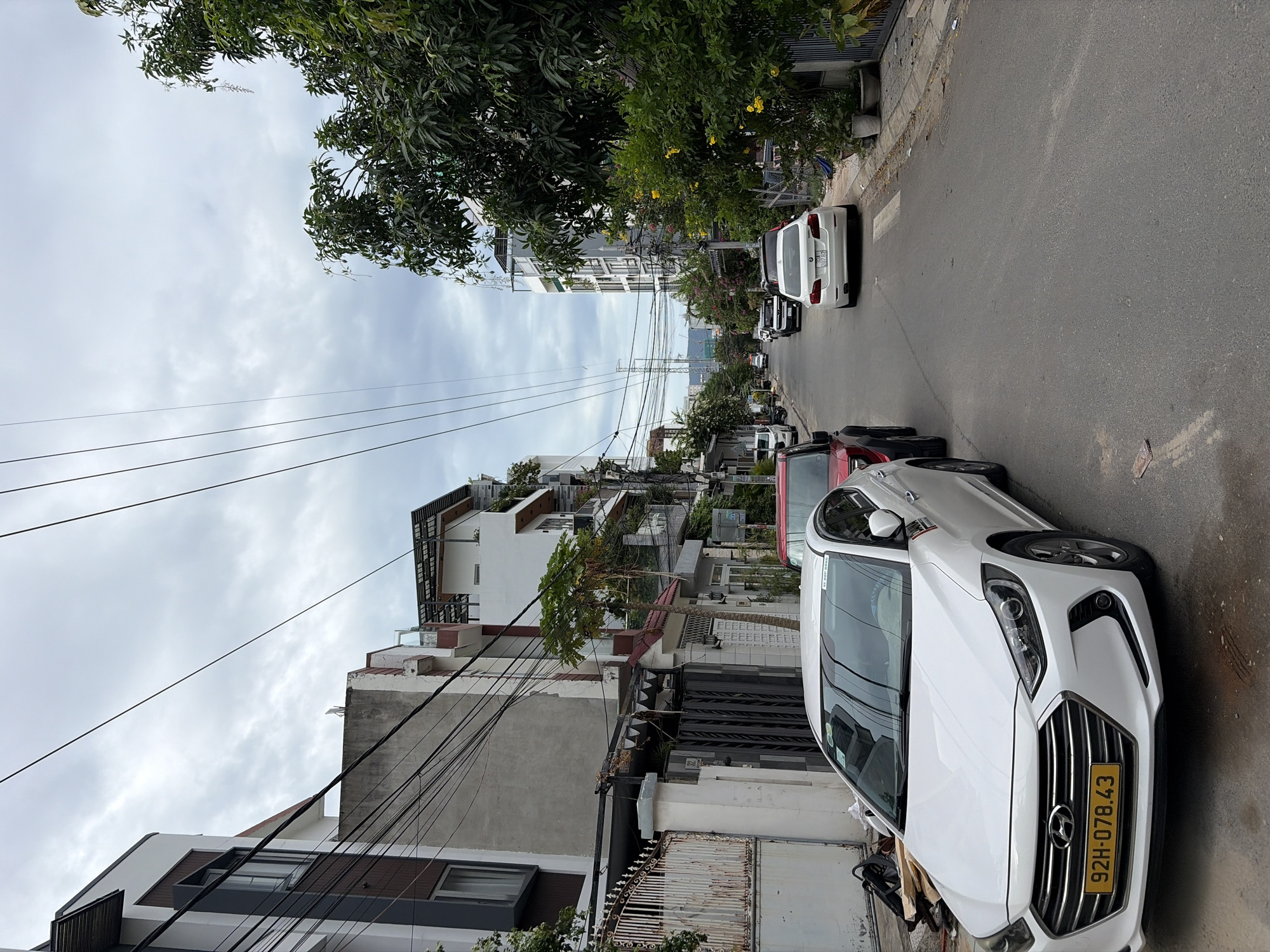 This is a street scene in Da Nang, Vietnam. In the foreground, a white Hyundai Elantra is parked on the left side of the road. Its license plate reads "92H-078.43". Behind it, a red SUV is also parked. The street is lined with multi-story buildings on both sides, some with balconies and trees. Overhead, a dense network of electrical wires crisscrosses the sky. In the distance, taller modern buildings are visible, suggesting a mix of older and newer construction. The sky is overcast, with a grey, cloudy appearance. The overall impression is an urban street with typical city infrastructure and traffic.