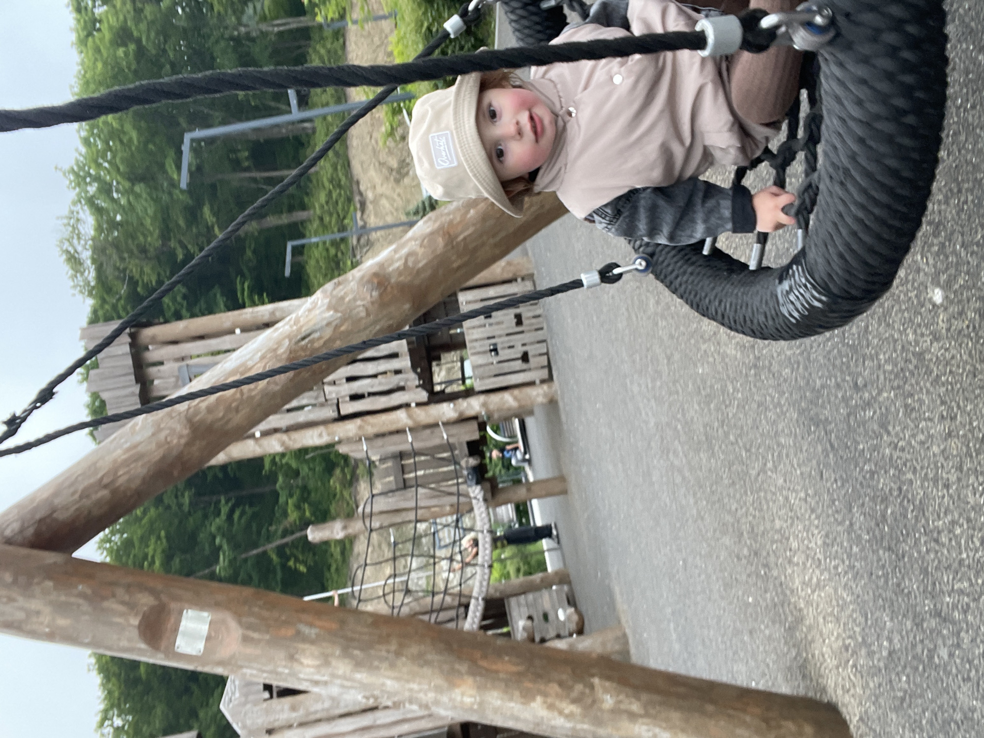 A young child is sitting in a large, black, circular rope swing at a playground. The child is wearing a light-colored bucket hat, a beige vest over a gray and black jacket, and brown pants. The child's left hand is resting on the rope of the swing. In the background, there is a wooden playground structure with climbing nets and slides. Trees and greenery are visible behind the playground. The ground is covered in dark gray gravel. The scene appears to be an outdoor park or playground. There is no indication of the city of Vladivostok, Russia.