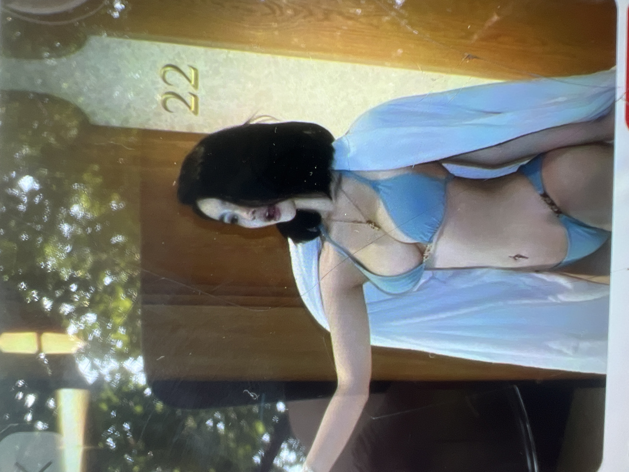 In the city of Kara-Dıykan, Kyrgyzstan, a young woman is depicted wearing a light blue bikini and a white sheer wrap. She has dark hair and is looking to her right. Behind her, a pillar displays the number "22". The environment appears to be outdoors with foliage visible in the background.