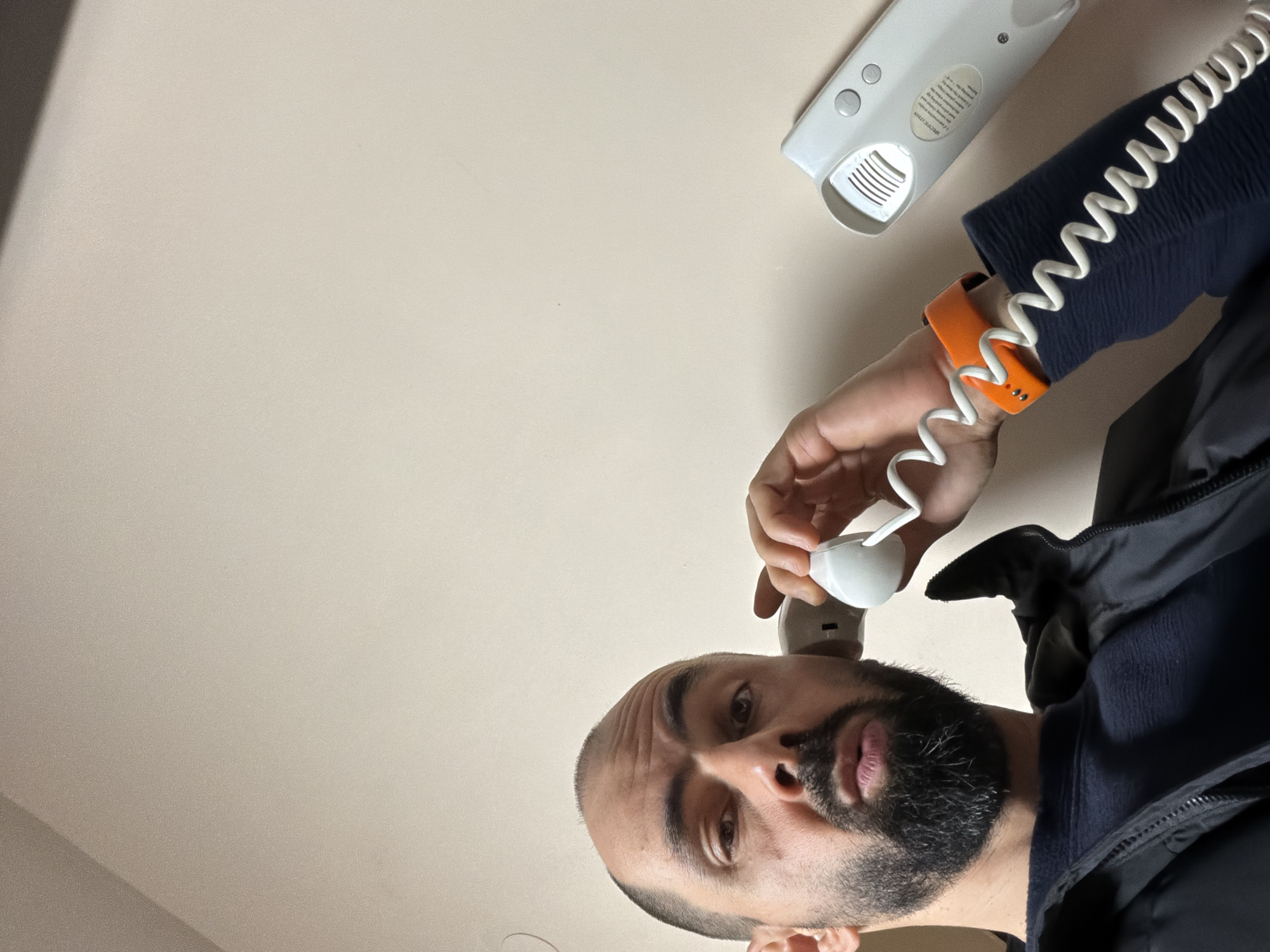 A low-angle shot depicts a man with dark hair, a beard, and an orange wristband. He is holding a white telephone receiver to his ear. The telephone base is partially visible in the upper right corner of the frame. The man is wearing a dark blue long-sleeved shirt and a dark jacket. The background appears to be a plain, light-colored ceiling or wall. The location is unknown and is not possible to tell from this image if it is in Plovdiv, Bulgaria.