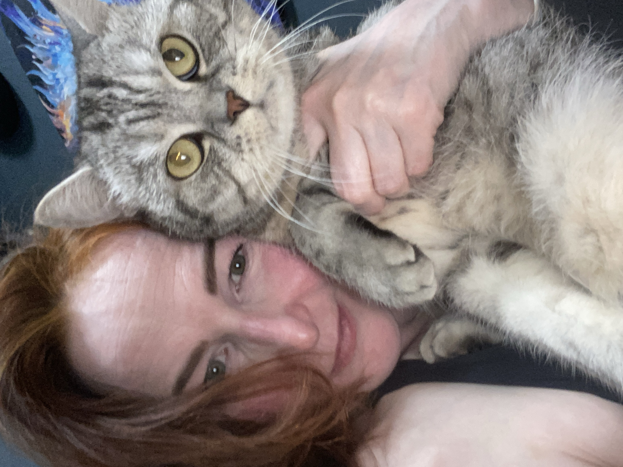 A woman with red hair is lying down, her head tilted towards the camera. A tabby cat with green eyes is resting on her face and chest, with a hand holding the cat. The background is a dark, indistinct surface. No information suggests that this scene is in Chaikovsky, Russia.