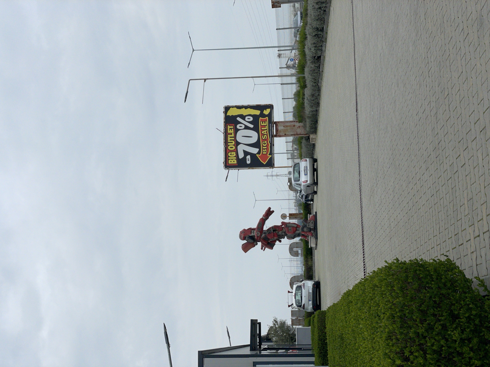 The image depicts a roadside scene in what appears to be the city of Asenovgrad, Bulgaria, identifiable by the presence of a large "BIG OUTLET -70% BIG SALE" sign. A large, reddish, humanoid robot statue is displayed along the roadside. Several parked vehicles are visible next to a sidewalk paved with interlocking concrete blocks and separated from the road by a shrubbery hedge. Streetlights are positioned along the road. The sky is overcast. A low-rise building with a flat roof is located near the robot statue.