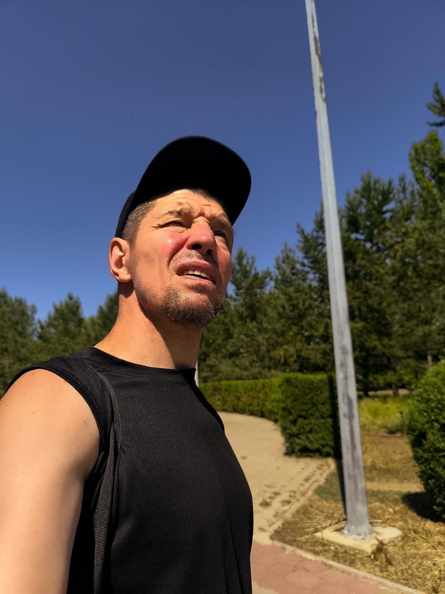 A man wearing a black sleeveless shirt and a black baseball cap is looking up at a bright blue sky. He has a short beard and stubble. His face is flushed, suggesting he may have been in the sun. He is outdoors, with green trees and bushes in the background. A tall, slender pole, possibly a light pole or flagpole, is visible to his right. The ground appears to be paved with red brick and dirt paths. The scene is likely in a park or public outdoor space in Qarağandı city administration, Kazakhstan.