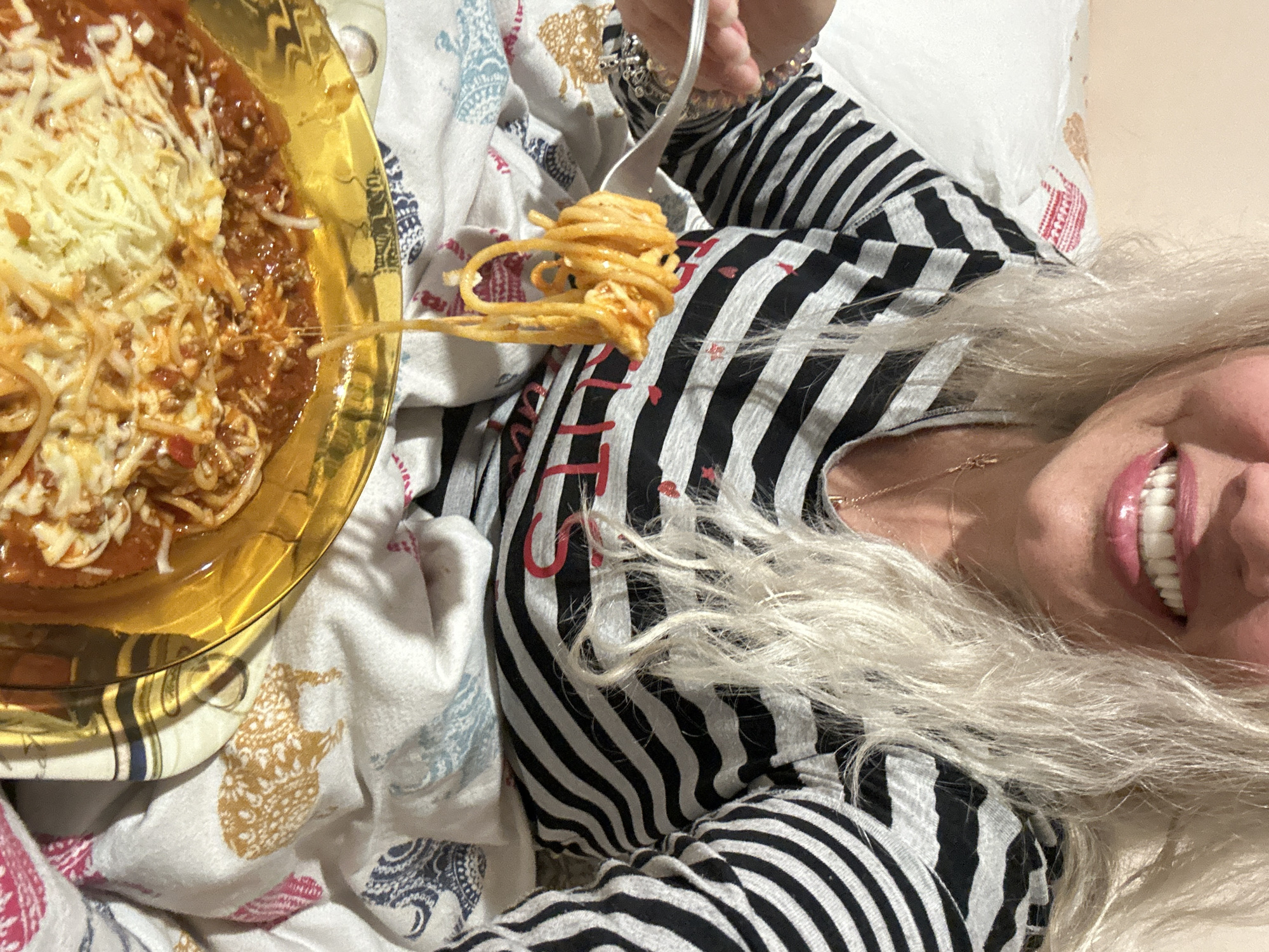The image depicts a woman with long, light-colored hair lying in bed. She is smiling, revealing white teeth. She wears a black and white striped shirt. A silver fork is raised with spaghetti twirled around it, positioned above her chest. To the left, there is a golden plate piled with spaghetti, meat sauce, and shredded cheese. The bed is covered with a patterned white sheet. The specific location within Sofia, Bulgaria is not discernible from the image.