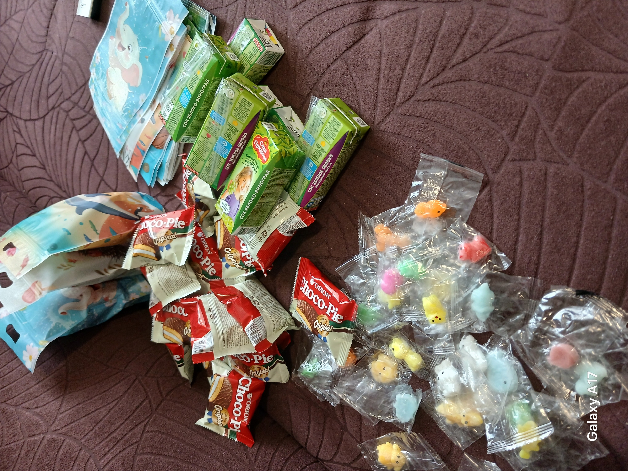 The image shows an assortment of children's items scattered on a brown, patterned surface. These include: several small juice boxes labeled "СОК ЯБЛОКО-ВИНОГРАД" and "СОК ЗЕЛЁНОЕ ЯБЛОКО," packages of "Choco-Pie" snacks, small plastic bags with miniature animal-shaped toys, and bags with cartoon elephant designs. The "Galaxy A17" watermark suggests the image was taken with that specific phone model. The presence of "Сады Придонья" juice boxes and the Cyrillic text suggests the location is in a Russian-speaking area, potentially Yekaterinburg, Russia, but without further information, it is impossible to confirm the exact location.